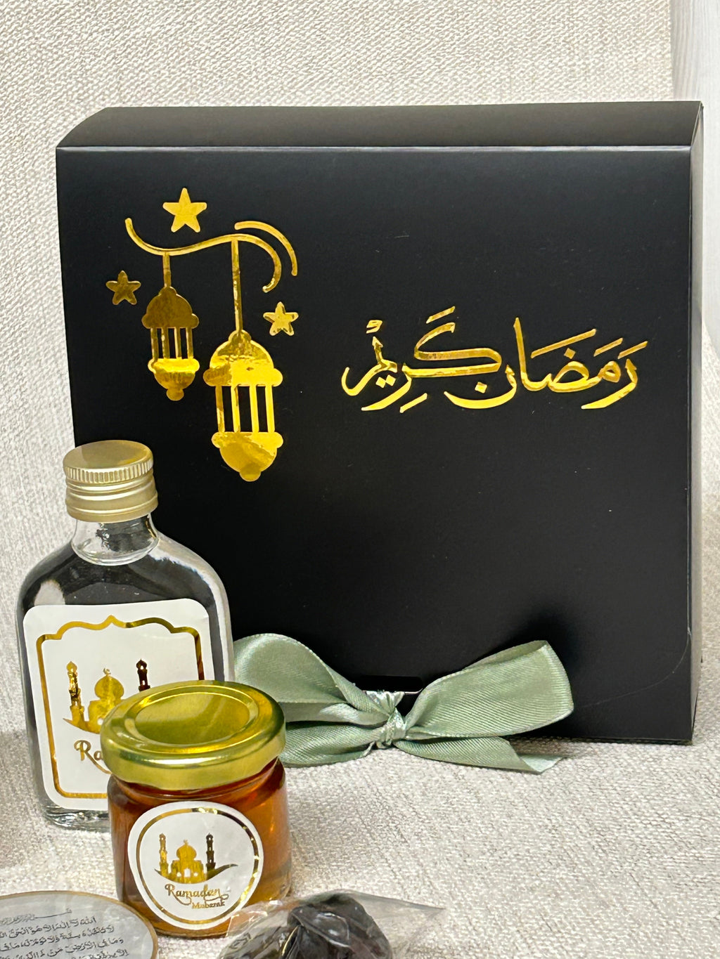 Gift set with black box with gold Ramadan Kareem text and a sage green ribbon, Zamzam flask, and mini honey jar on a textured surface