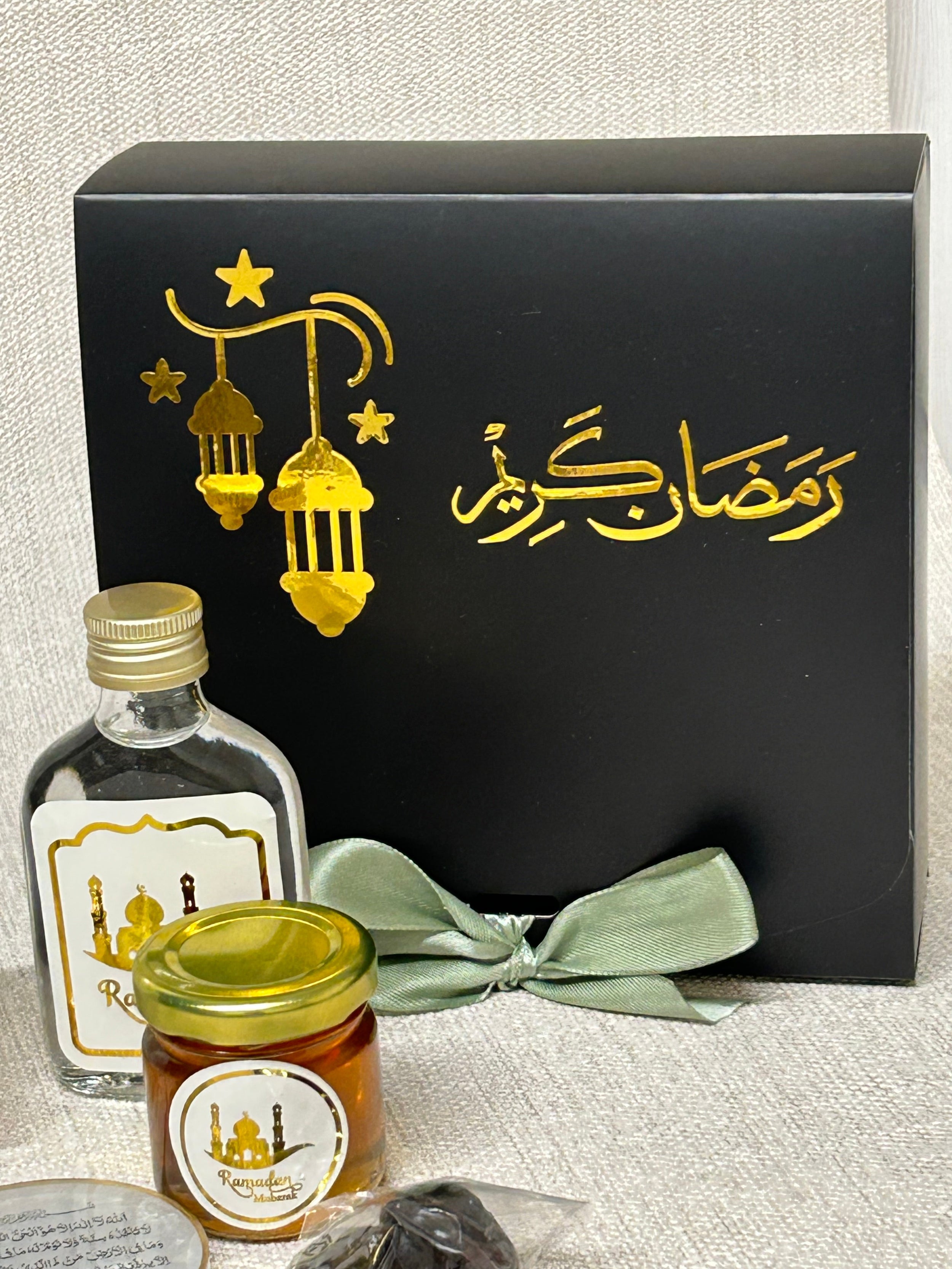 Gift set with black box with gold Ramadan Kareem text and a sage green ribbon, Zamzam flask, and mini honey jar on a textured surface