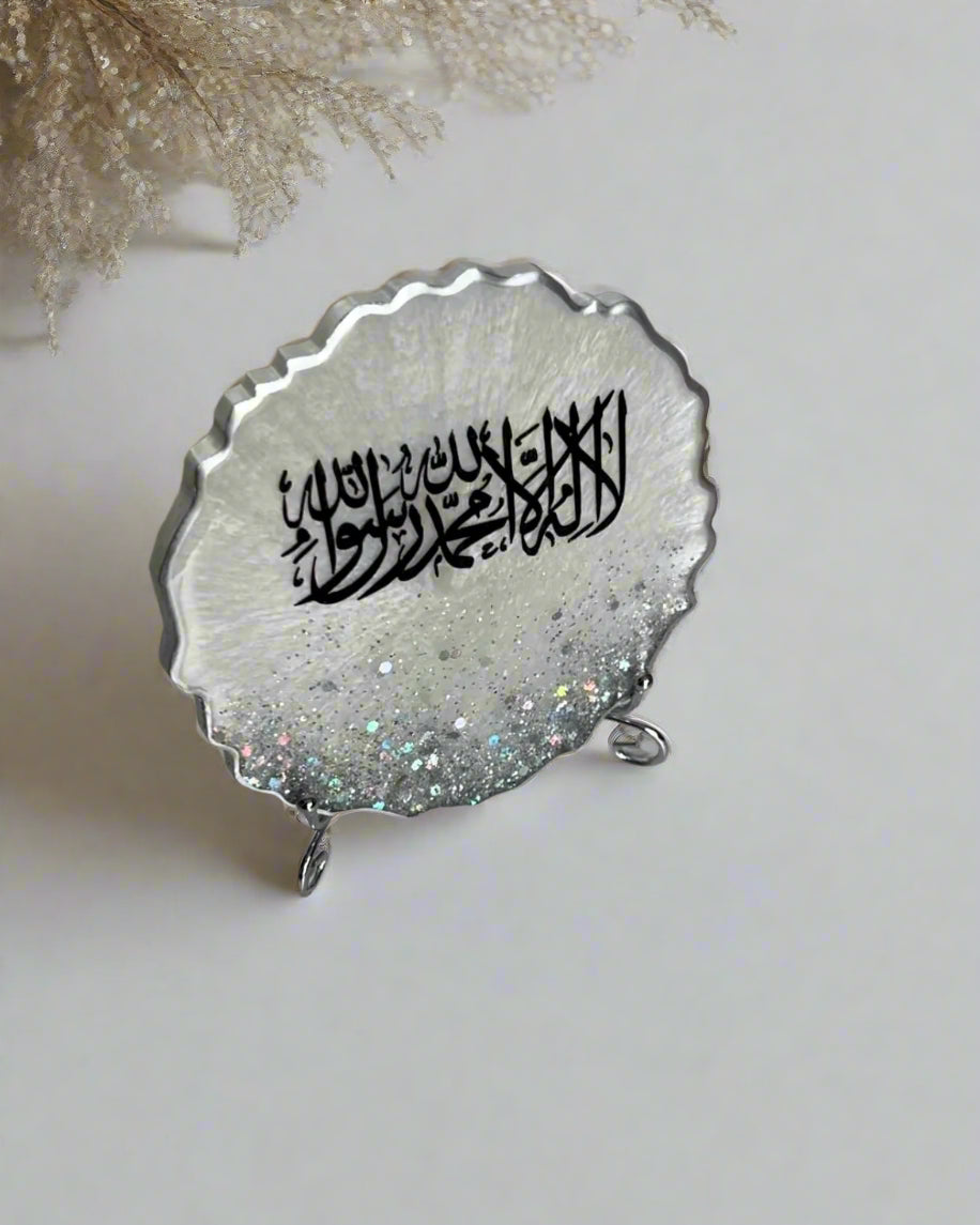 A silver resin plaque with Arabic calligraphy, featuring shahada, placed on a table with decorative items around it.