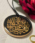 Round black pendant with gold Arabic calligraphy on a white surface with a red flower.