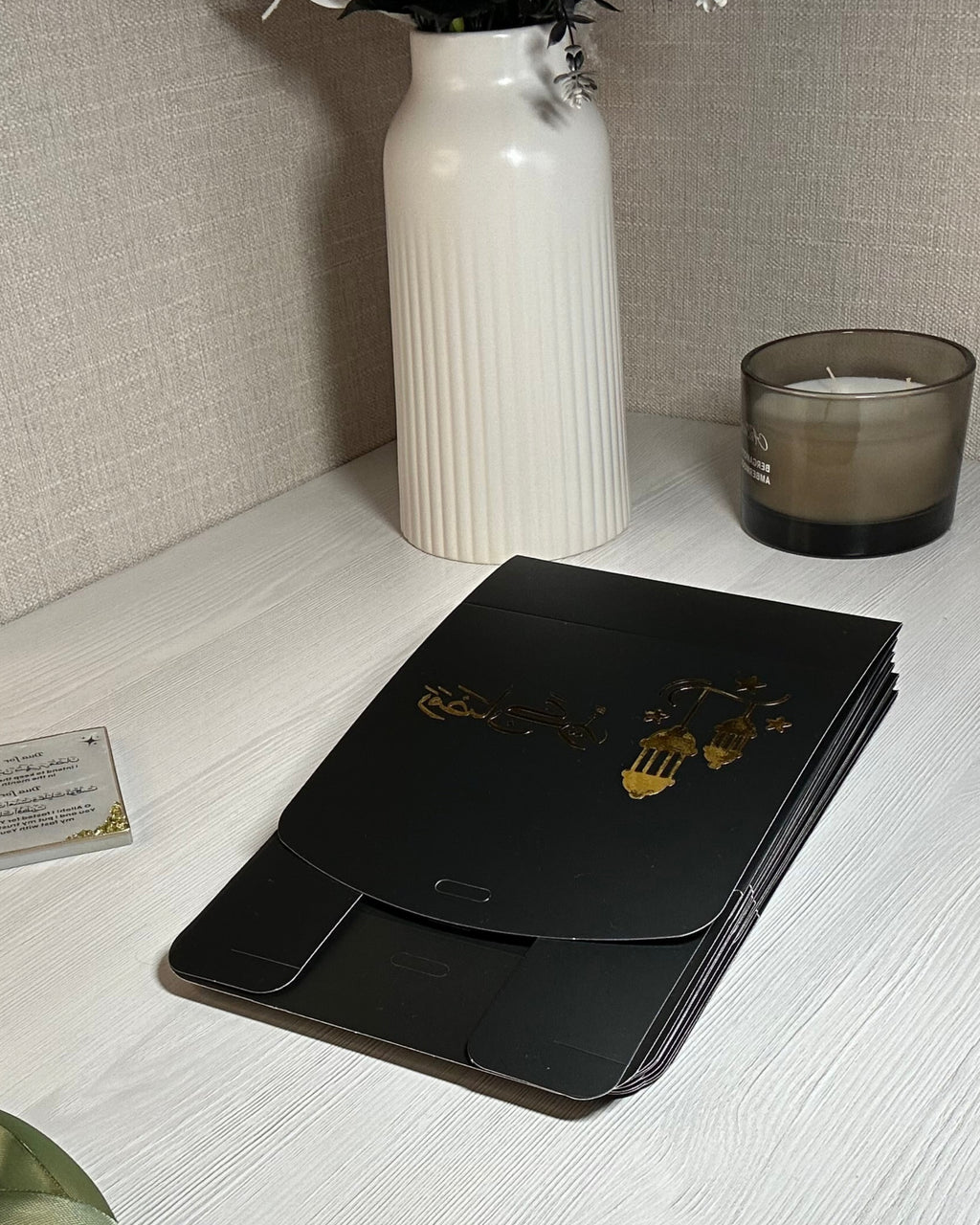 Black flat packed gift box with gold Ramadan Kareem design on a white surface with a vase and candle in the background.