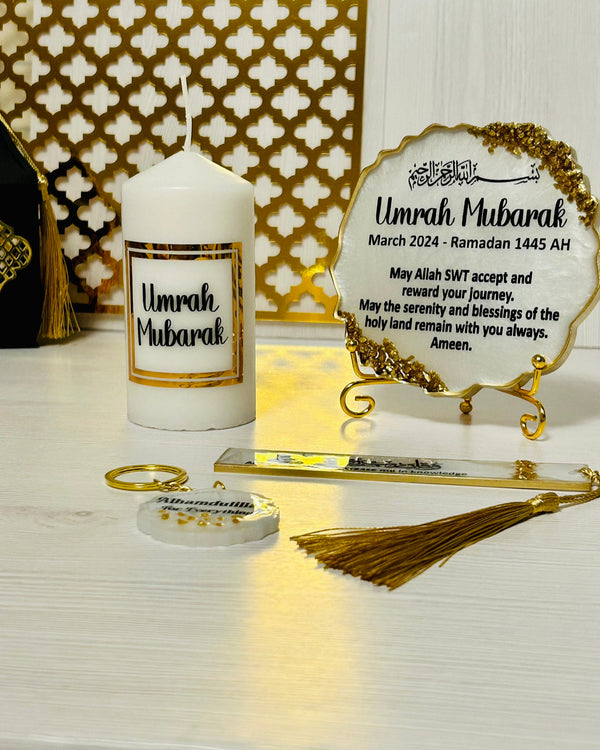 A gift set for Umrah, including a resin plaque with a gold crushed glass and a quote, a Quran bookmark, a keyring, chocolates, a candle, and an artificial flower, all displayed on a white surface with a decorative background.