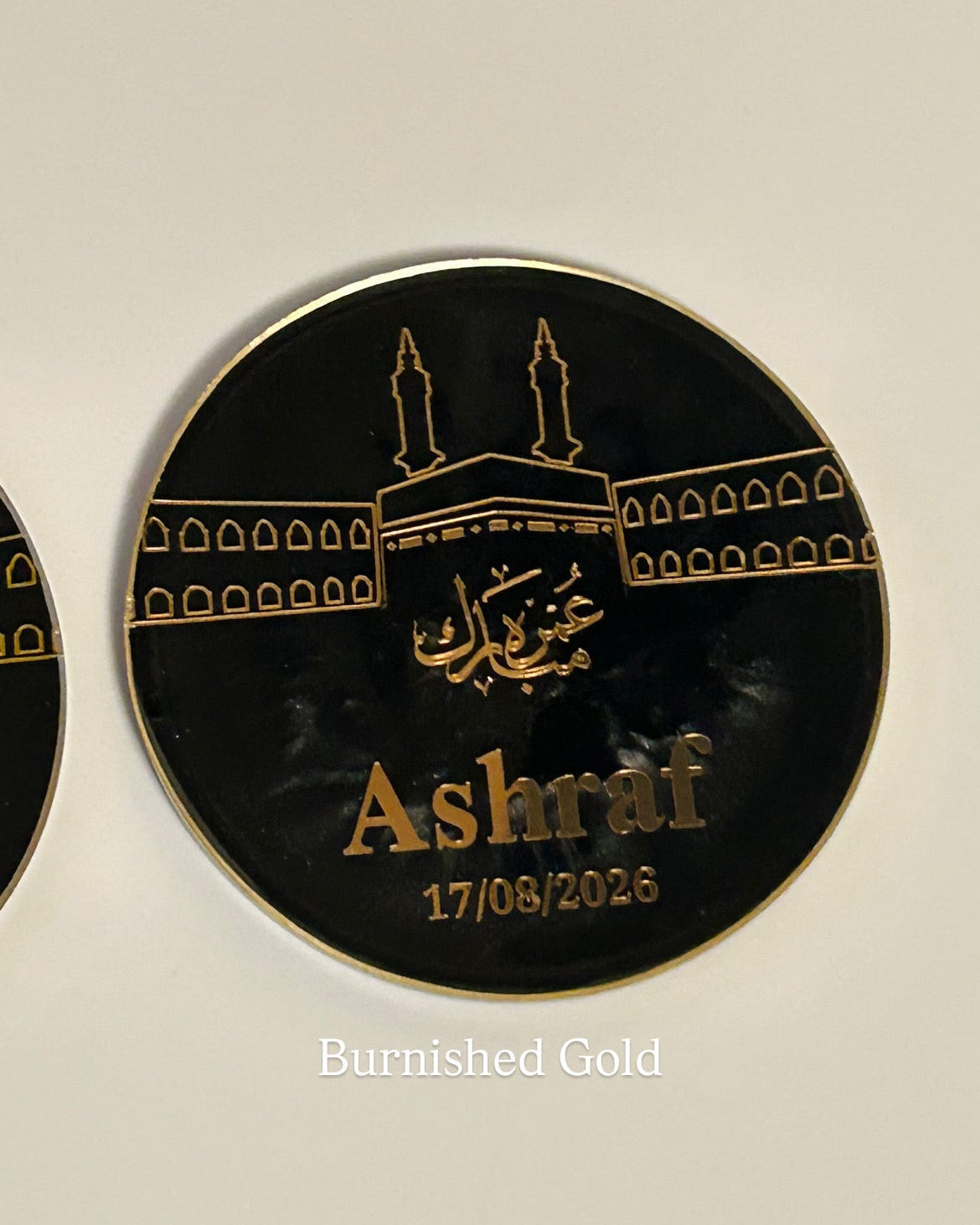 A round black personalised plaque featuring gold Islamic calligraphy and a mosque illustration, with the name 'Ashraf' and the date '17/08/2023' printed in gold, shown on a light background.