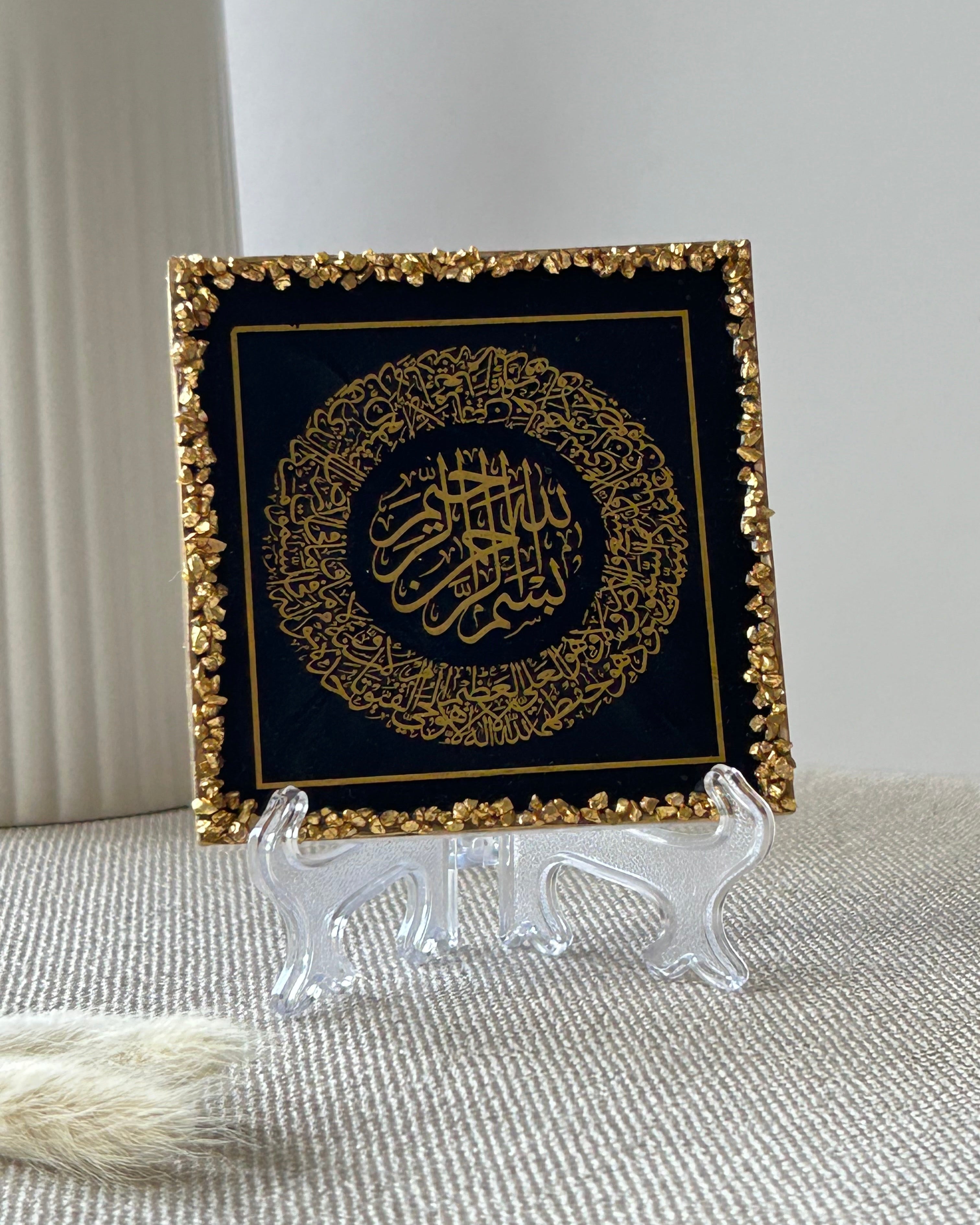 A square decorative plaque featuring gold Arabic calligraphy on a black background, bordered with textured gold crushed glass trim, displayed on a clear frosted plastic display stand on a beige textured surface.