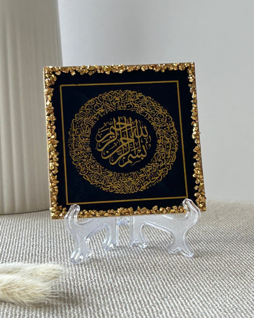 A square decorative plaque featuring gold Arabic calligraphy on a black background, bordered with textured gold crushed glass trim, displayed on a clear frosted plastic display stand on a beige textured surface.