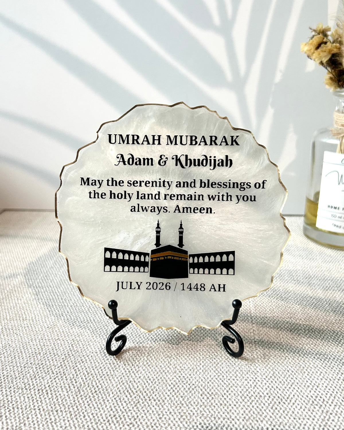 A white personalized round, scalloped-edge resin plaque, featuring black text and a black illustration of the Kaaba. The plaque is displayed on a small black metal stand on a textured beige fabric surface, with soft leaf shadows cast on a white wall behind it.
