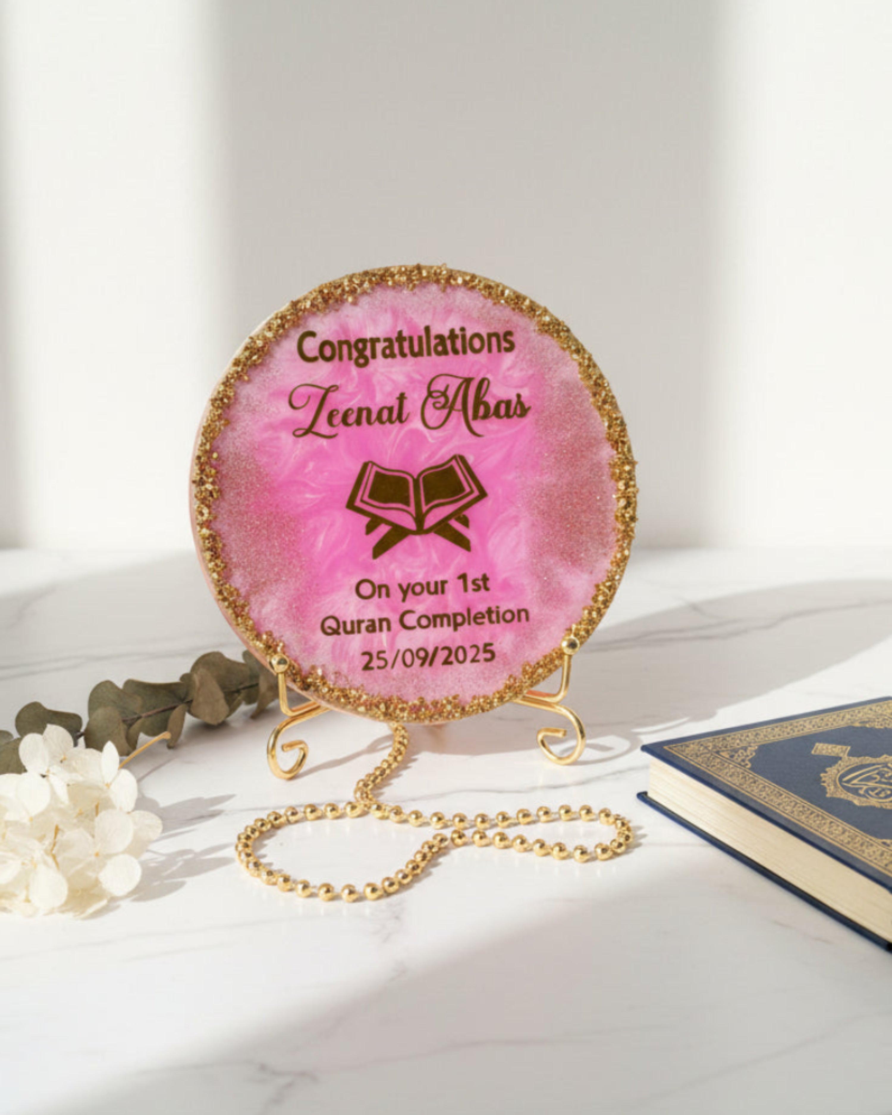 Pink Quran Completion decorative resin plaque with gold text and gold crushed glass around the edges resting on a gold metal display stand on a white background