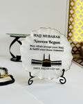 Pearlescent decorative Hajj Mubarak Plaque with text and the Kaaba and Mosque silhouette on a black display stand on a white background with decorative items