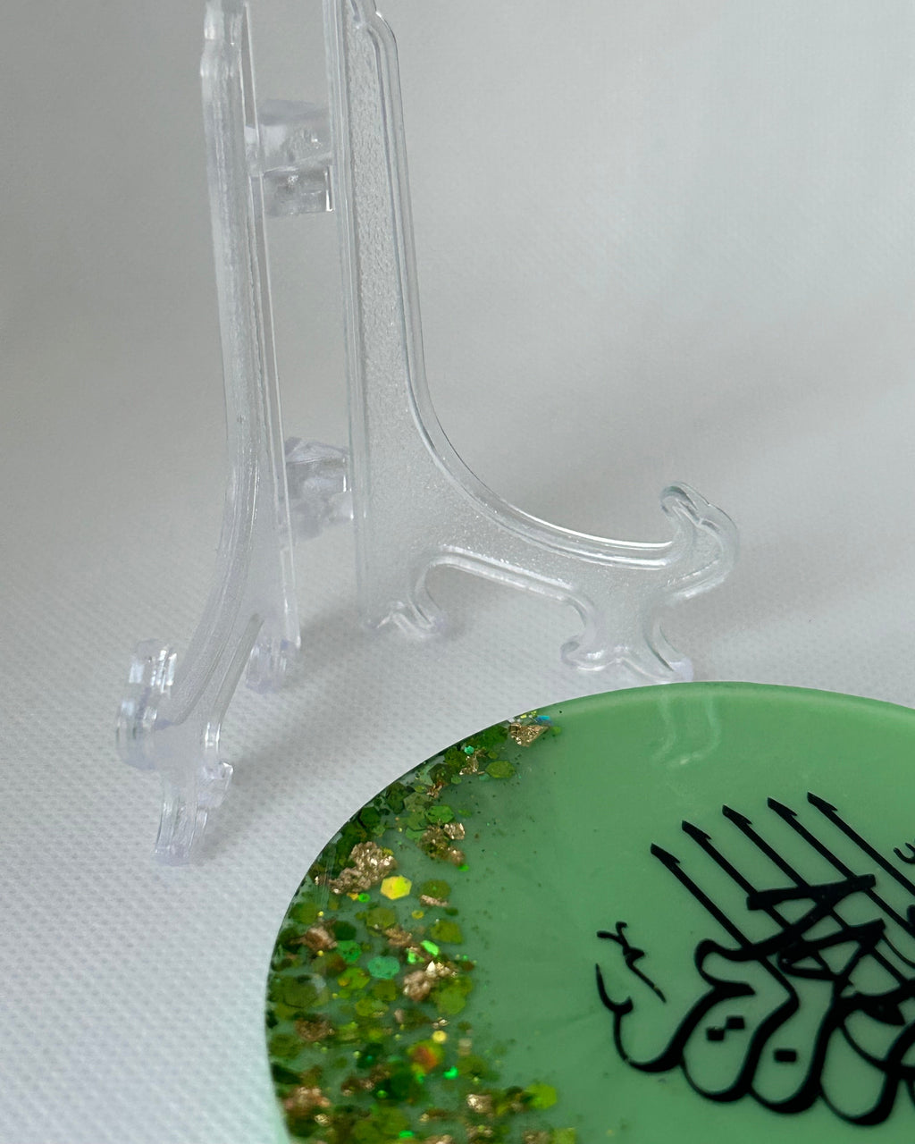 Green circular object with black Arabic calligraphy reading Bismillah Tasmiyah and gold shimmer glitter with gold leaf foiled placed sparsely displayed on a clear plastic display stand on a white background