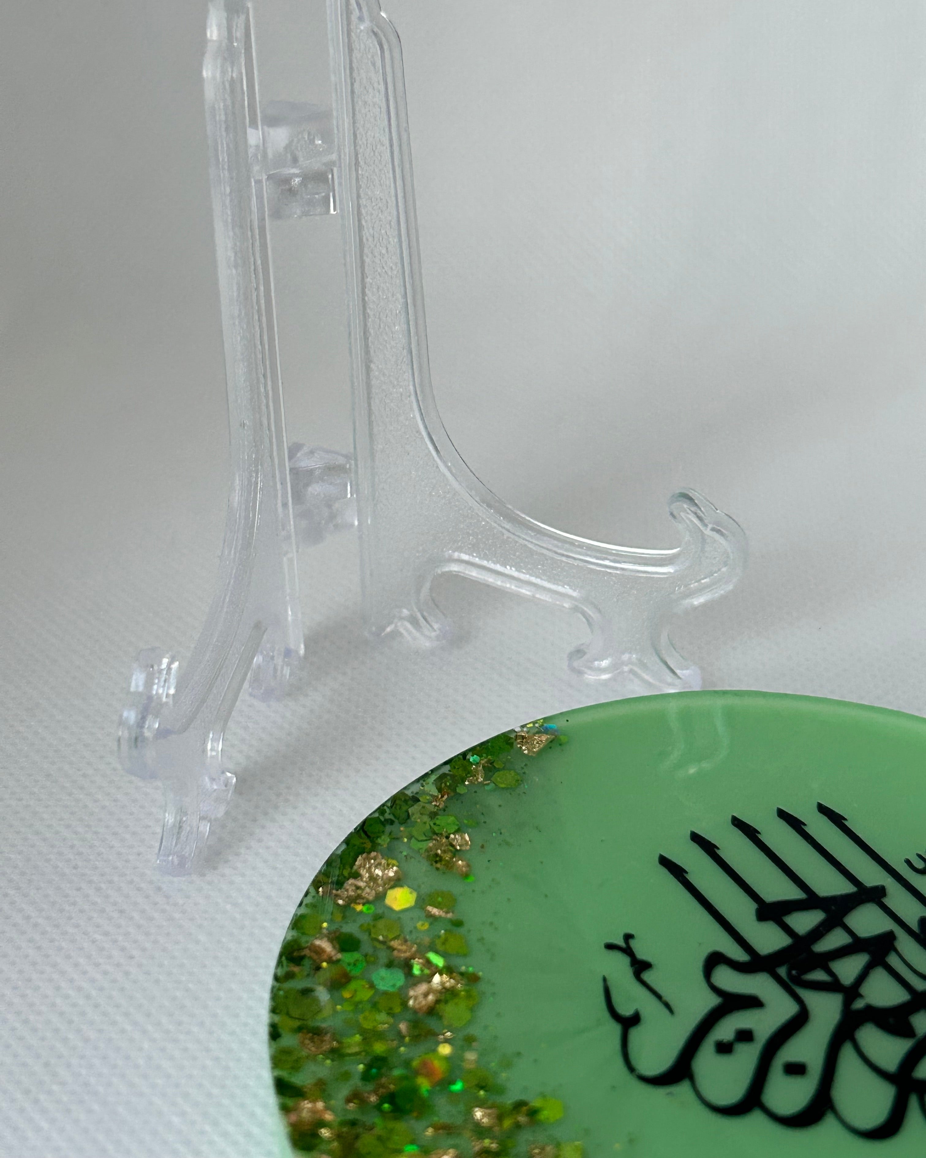 Green circular object with black Arabic calligraphy reading Bismillah Tasmiyah and gold shimmer glitter with gold leaf foiled placed sparsely displayed on a clear plastic display stand on a white background