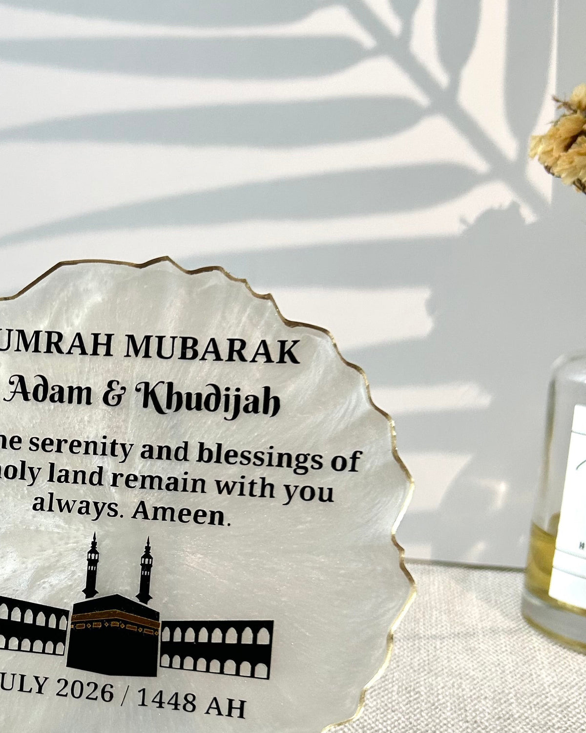 A personalized round white stone plaque with a gold-toned rim, displayed on a small black stand. The plaque features black printed text reading 'UMRAH MUBARAK Adam & Khudijah' and a black illustration of the Masjid.