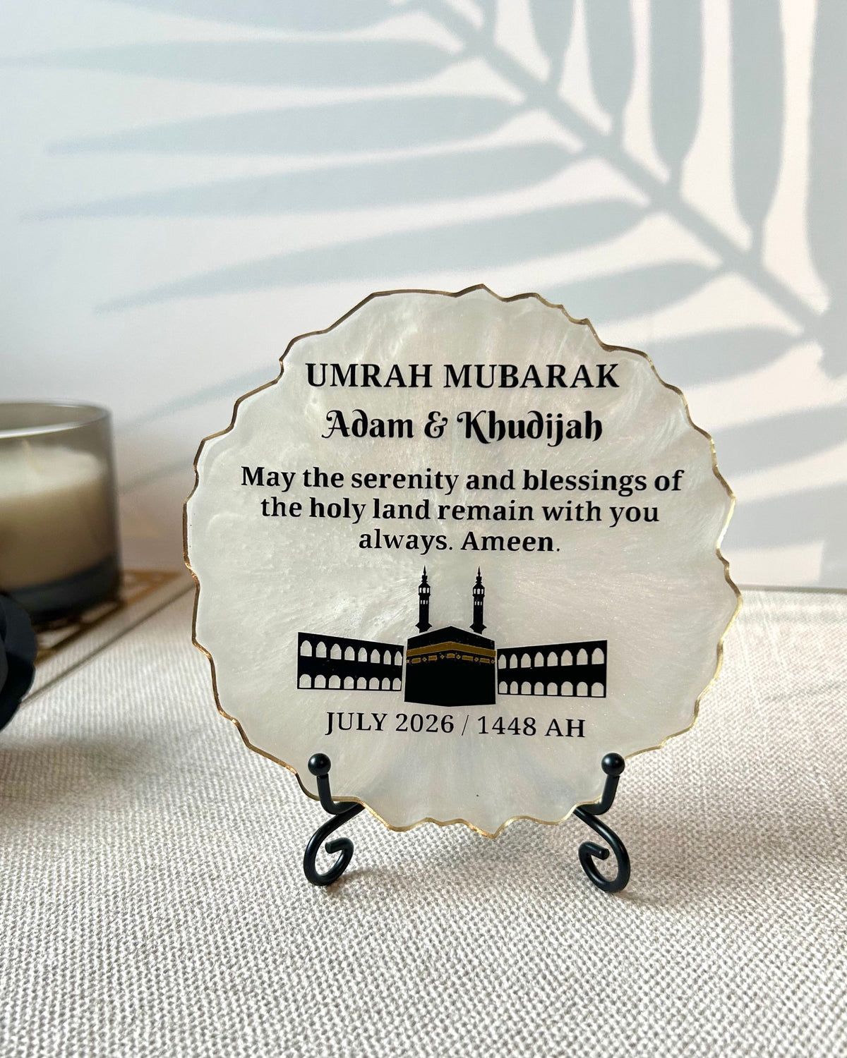 A personalised round plaque with a scalloped edge, featuring a black mosque illustration and the text 'UMRAH MUBARAK Adam & Khadijah' and a date, displayed on a small black stand on a textured fabric surface.