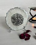 A silver resin plaque with intricate Arabic calligraphy, displayed on a stand, surrounded by rose petals on a wooden surface.