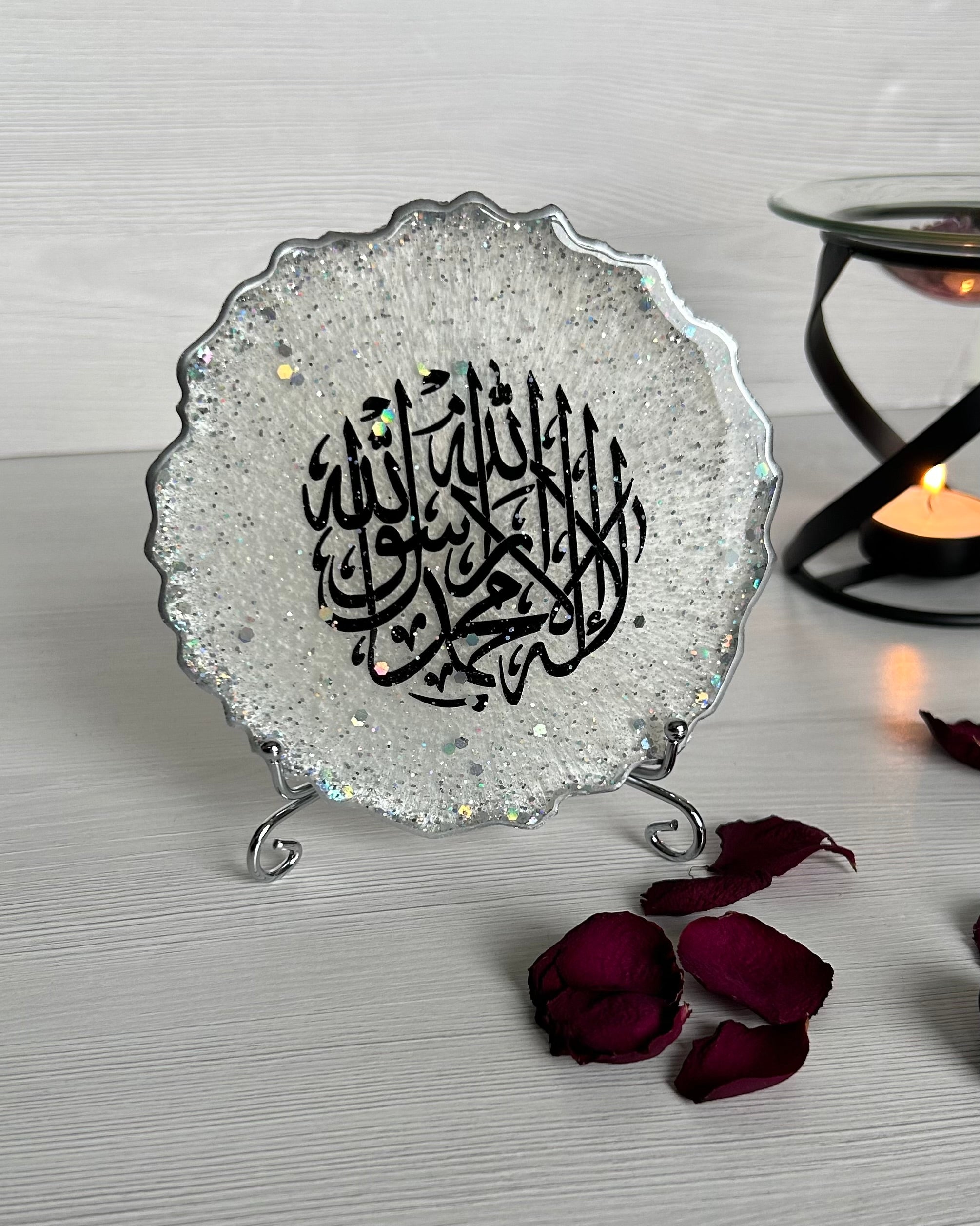A silver resin plaque with intricate Arabic calligraphy, displayed on a stand, surrounded by rose petals on a wooden surface.