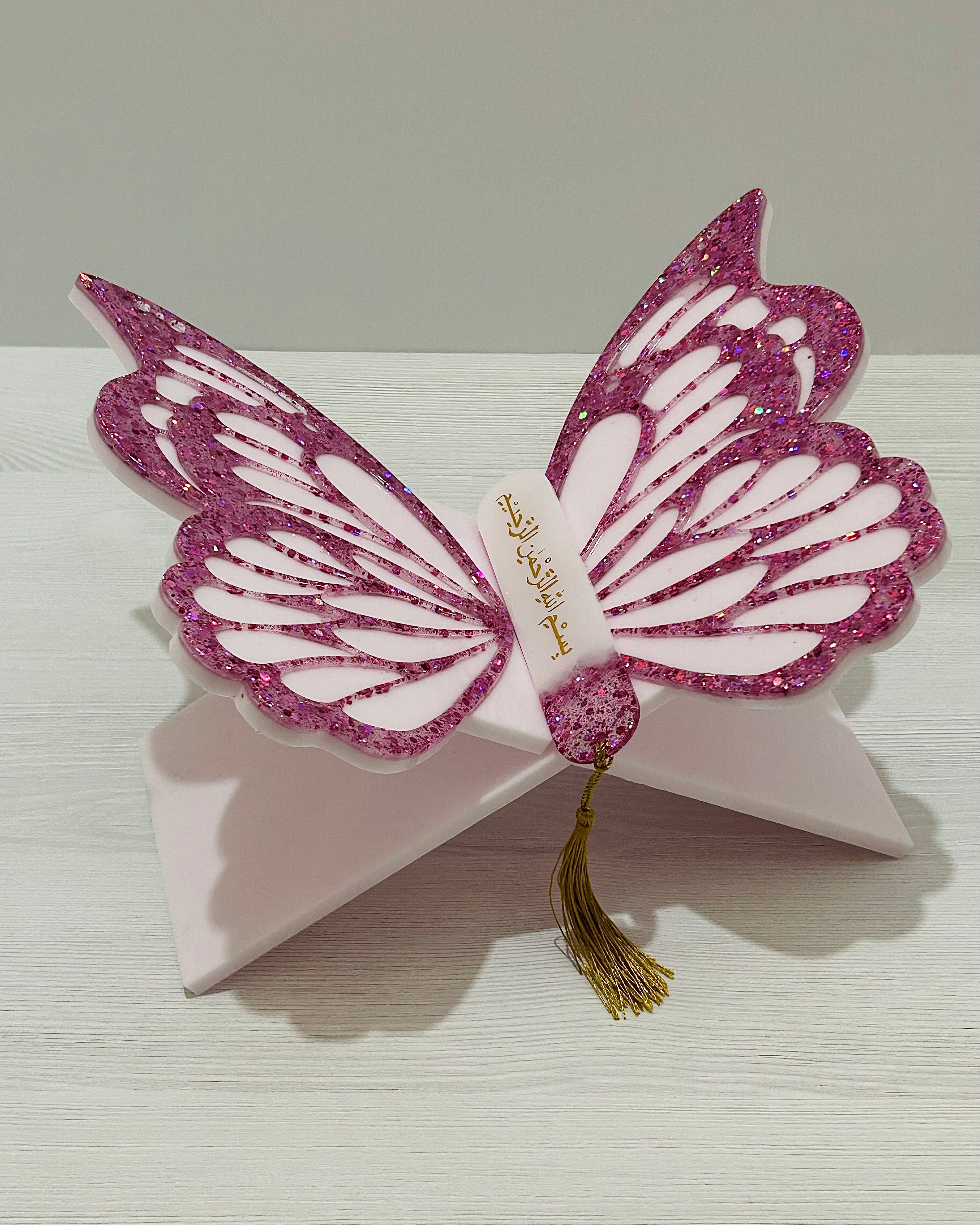 Pink butterfly-shaped Quran holder stand with wings decorated with pink glitter and matching bookmark on a white surface