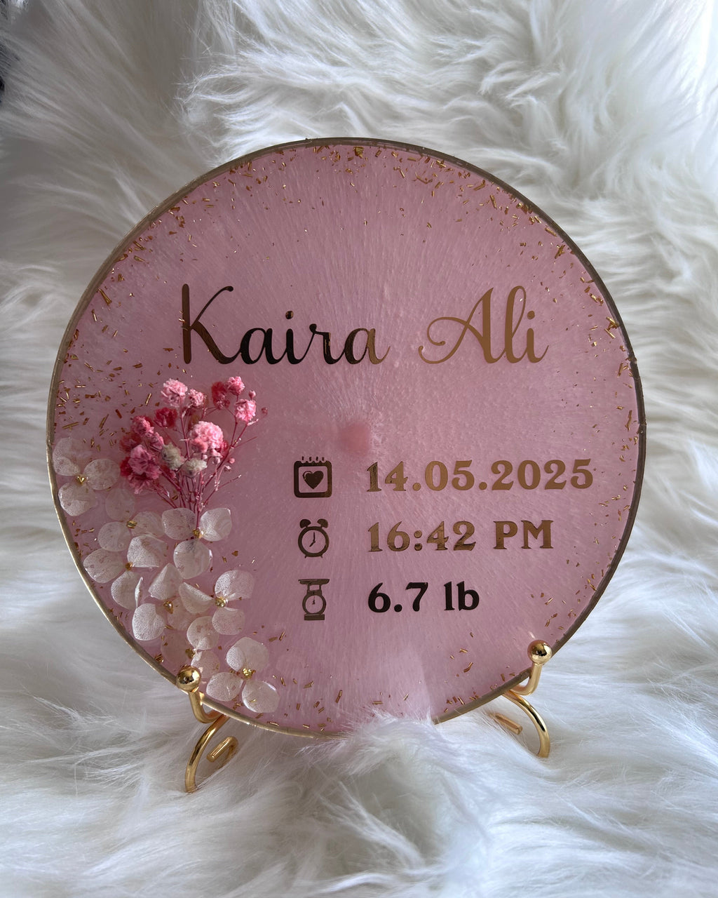 Round pink personalised resin baby announcement plaque with gold text on a white fluffy background