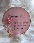 Round pink personalised resin baby announcement plaque with gold text on a white fluffy background