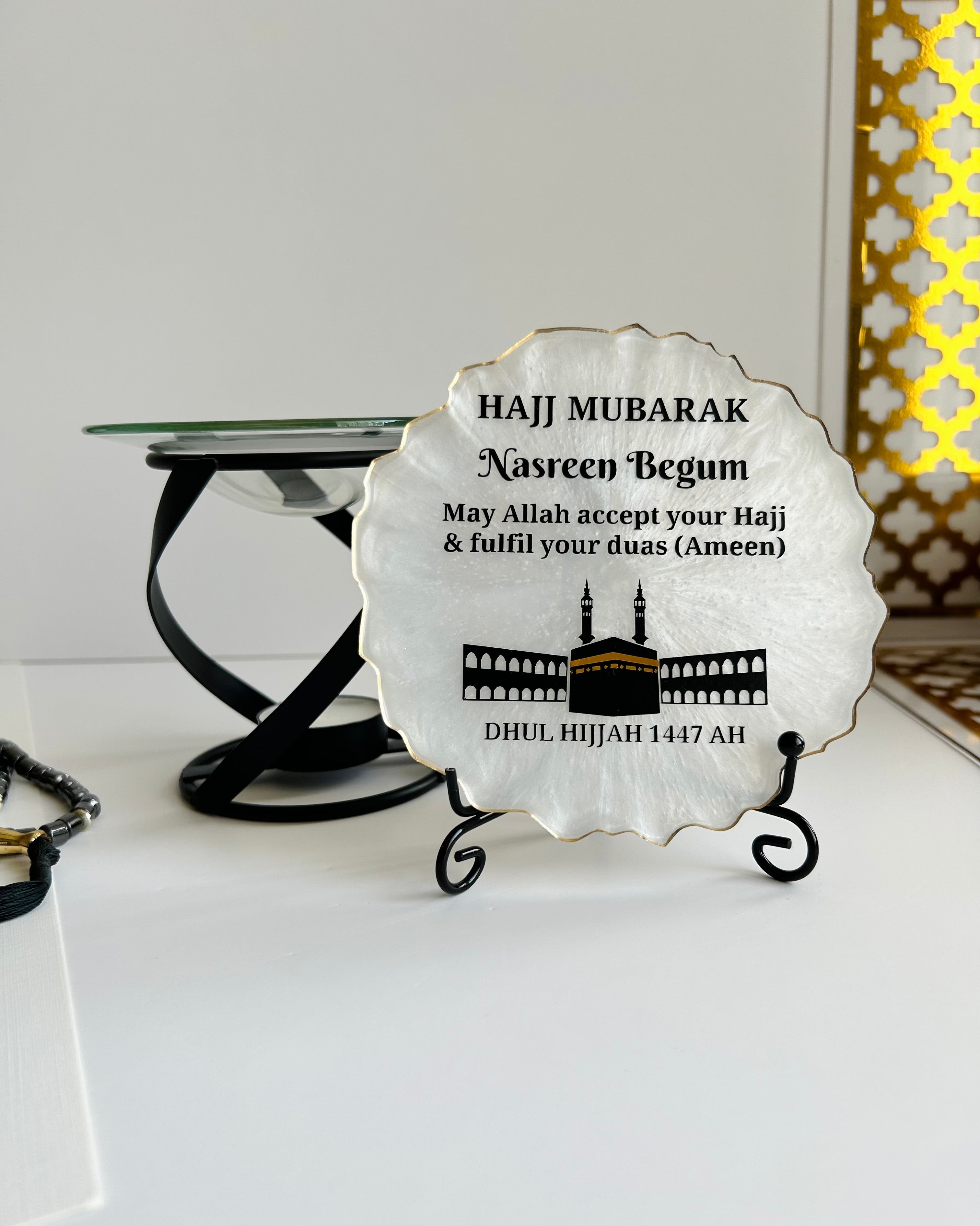 Pearlescent decorative Hajj Mubarak Plaque with text and the Kaaba and Mosque silhouette on a black display stand on a white background with decorative items