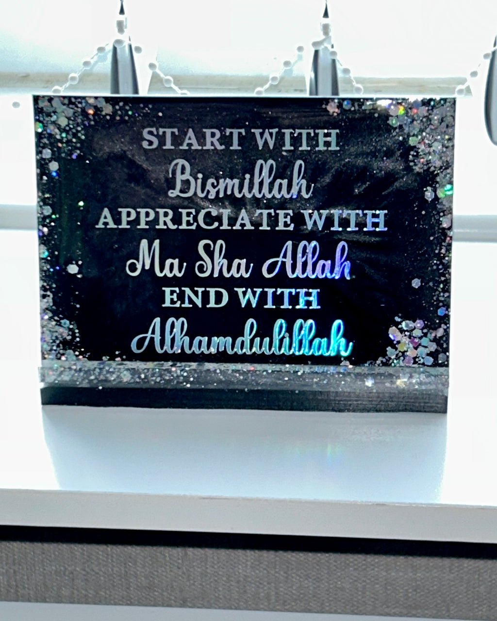 Decorative banner with Arabic text on a black background with glitter, hanging against a window.