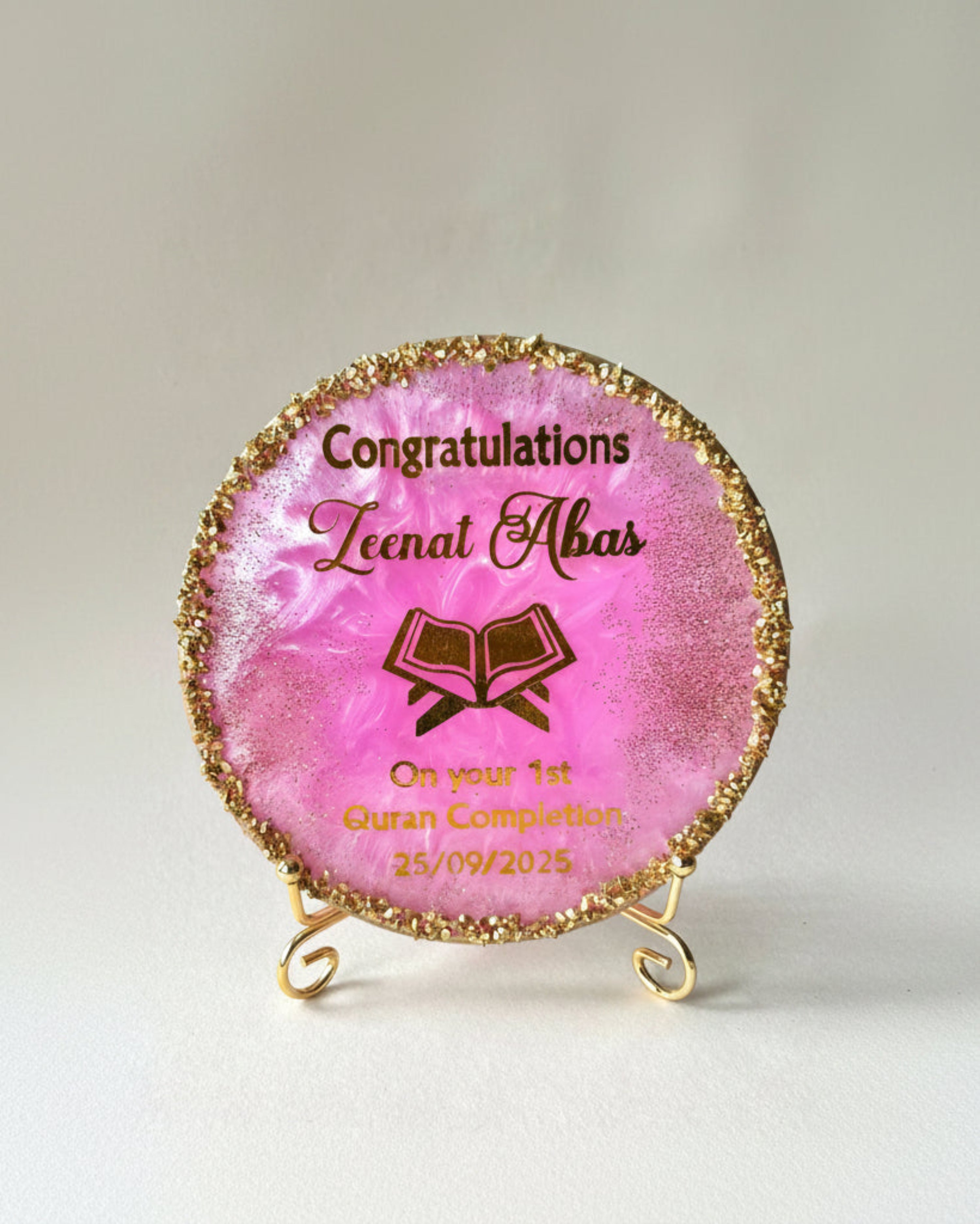 Pink Quran Completion decorative resin plaque with gold text and gold crushed glass around the edges resting on a gold metal display stand on a white background