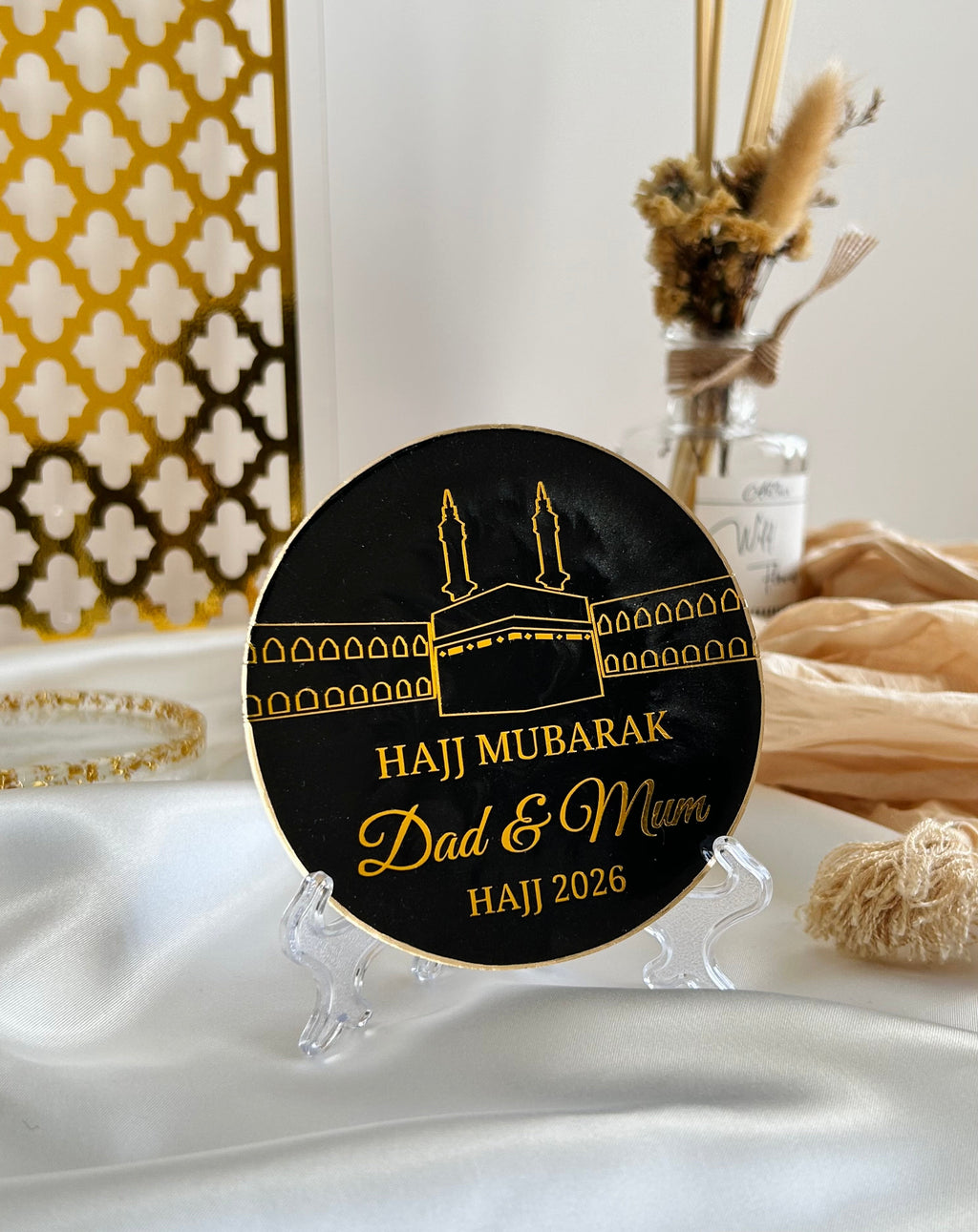 Decorative plate with 'Hajj Mubarak' design on a stand, featuring text and a religious symbol on a white satin background with decorative items