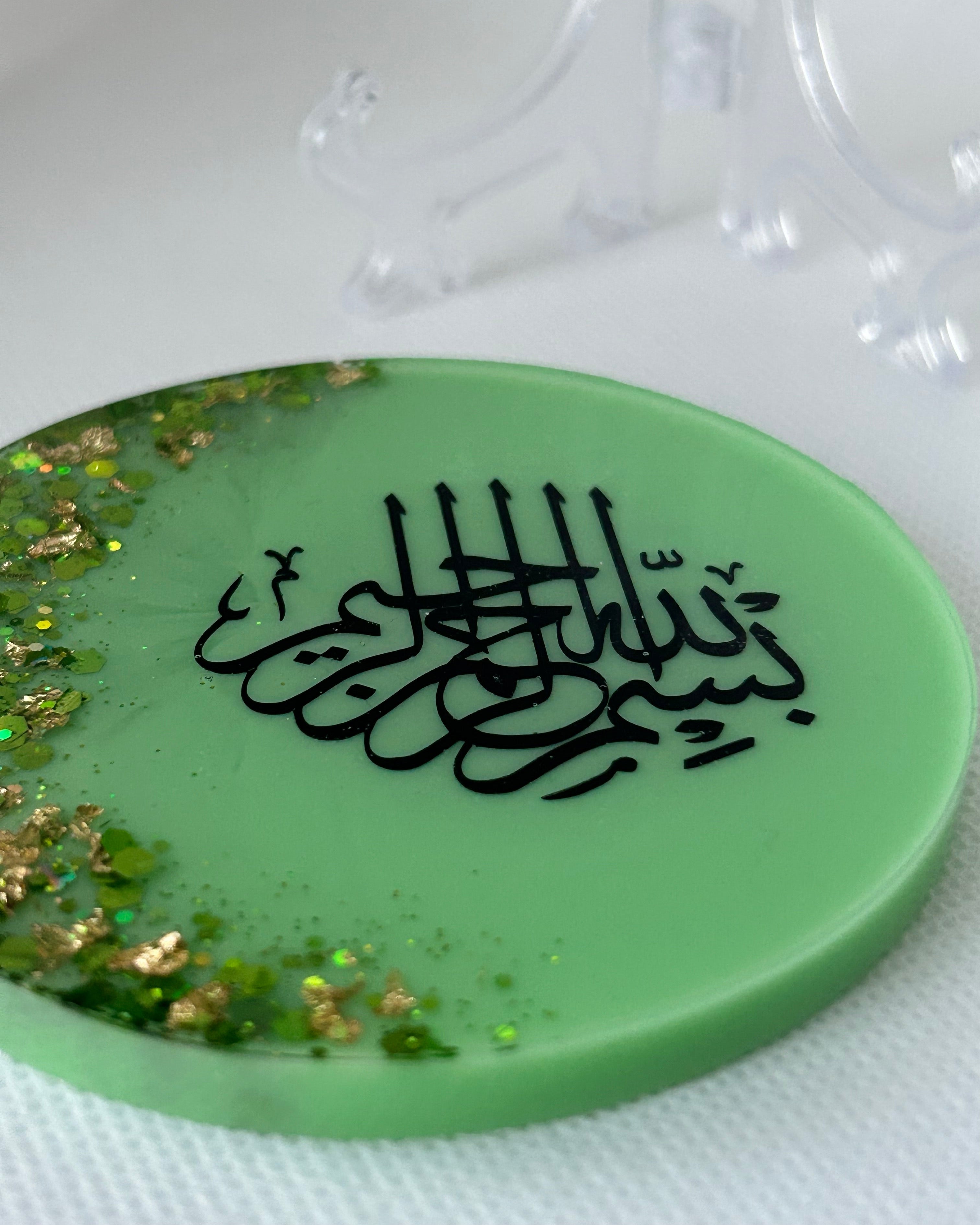 Green circular object with black Arabic calligraphy reading Bismillah Tasmiyah and gold shimmer glitter with gold leaf foiled placed sparsely displayed on a clear plastic display stand on a white background
