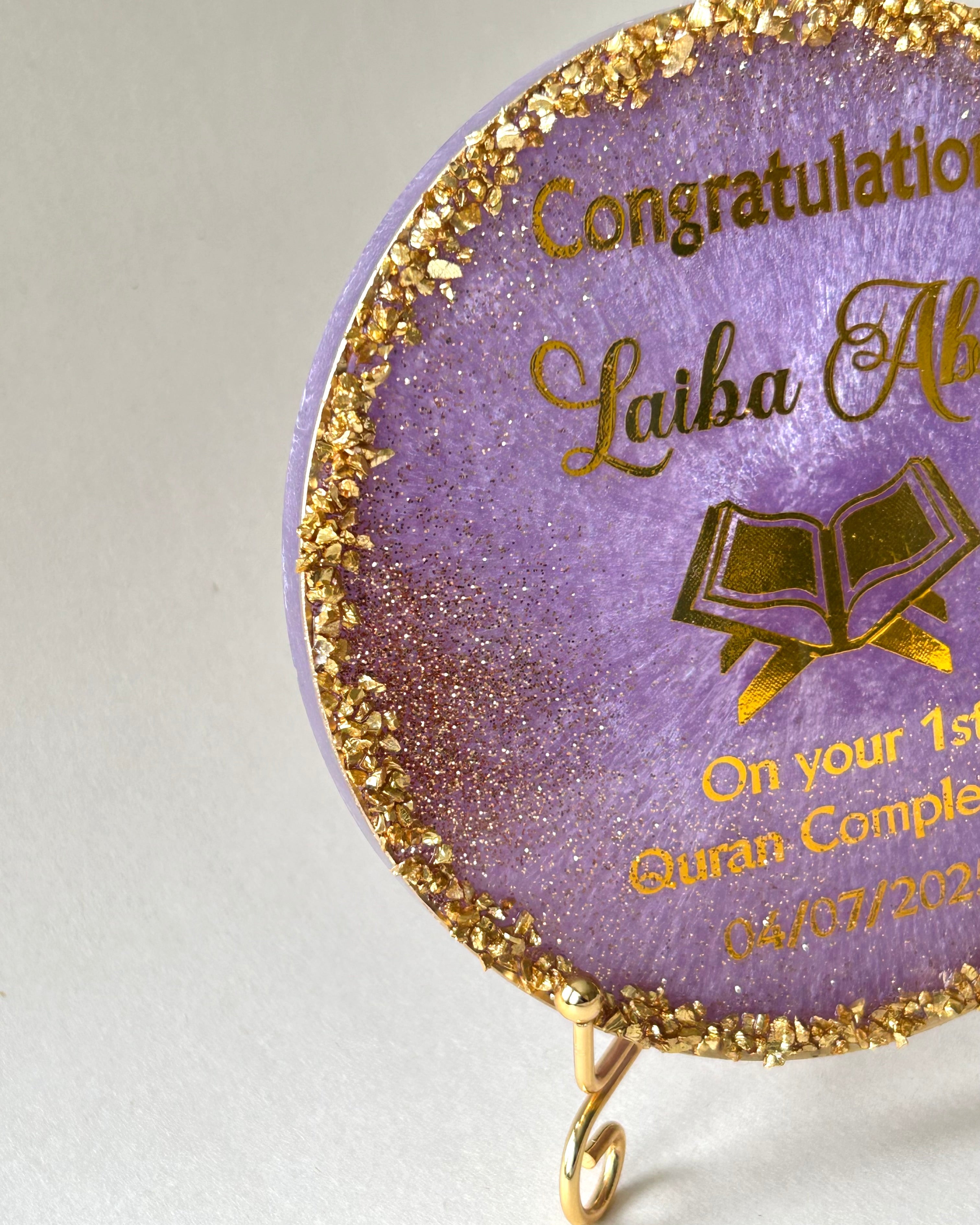 side of decorative Quran completion plaque with gold text, gold gliter, gold crushed glass and a Quran icon resting on a gold metal display stand on a white background