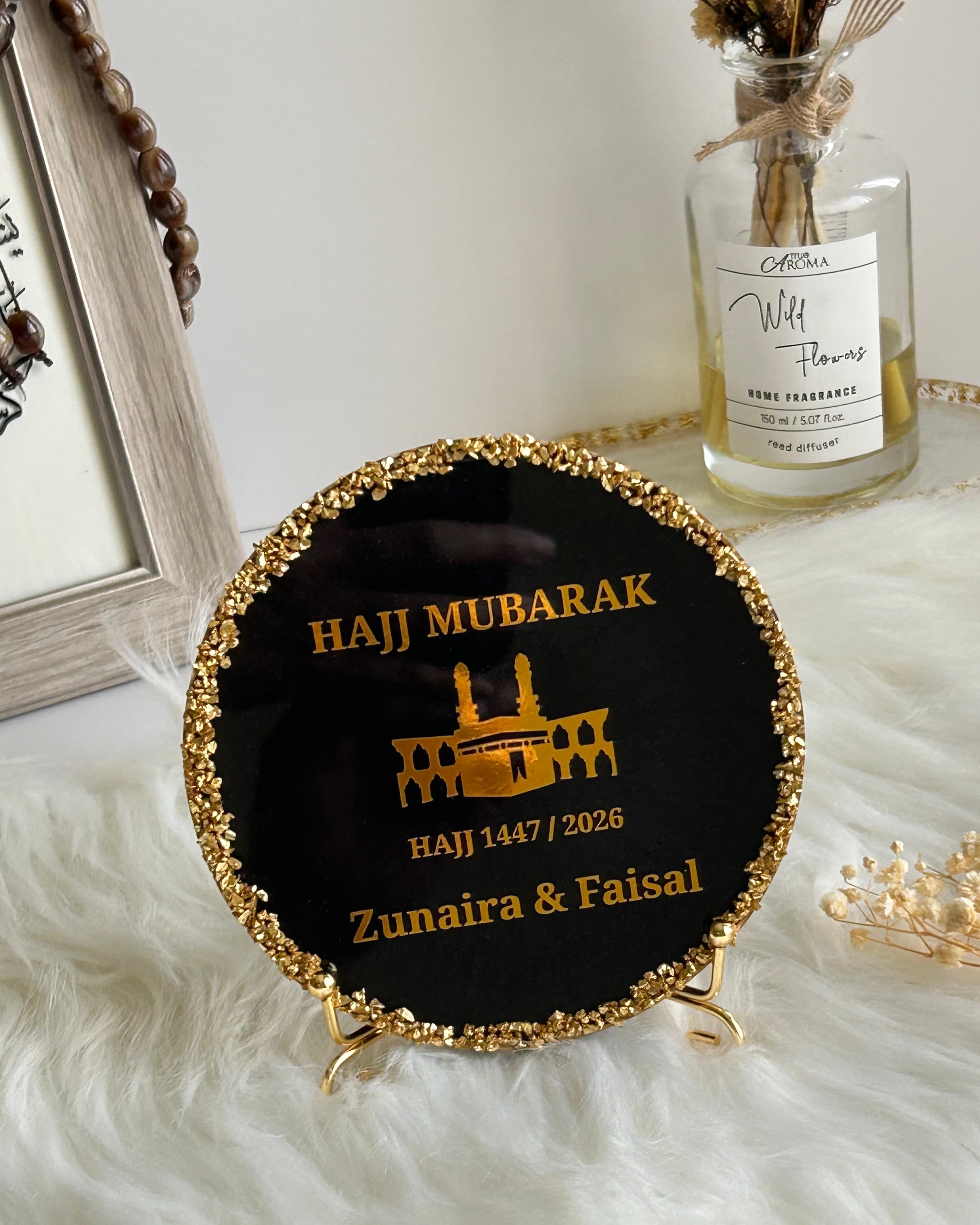 Round black decorative Hajj Mubarak plaque with gold rim and text on a white surface with decorative elements.