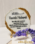white plate with gold decorative edges and black text reading the shahada in Arabic and Umrah Mubarak in English displayed on a gold display stand next to purple flowers