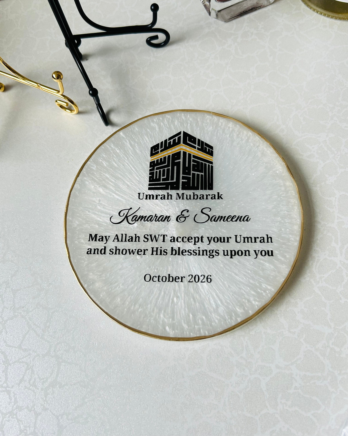 A round pearlescent white resin plaque with a gold rim, with a black Kaaba in Kufic style block reading Basmala and a personalised Umrah messaging, displayed on a light textured surface.