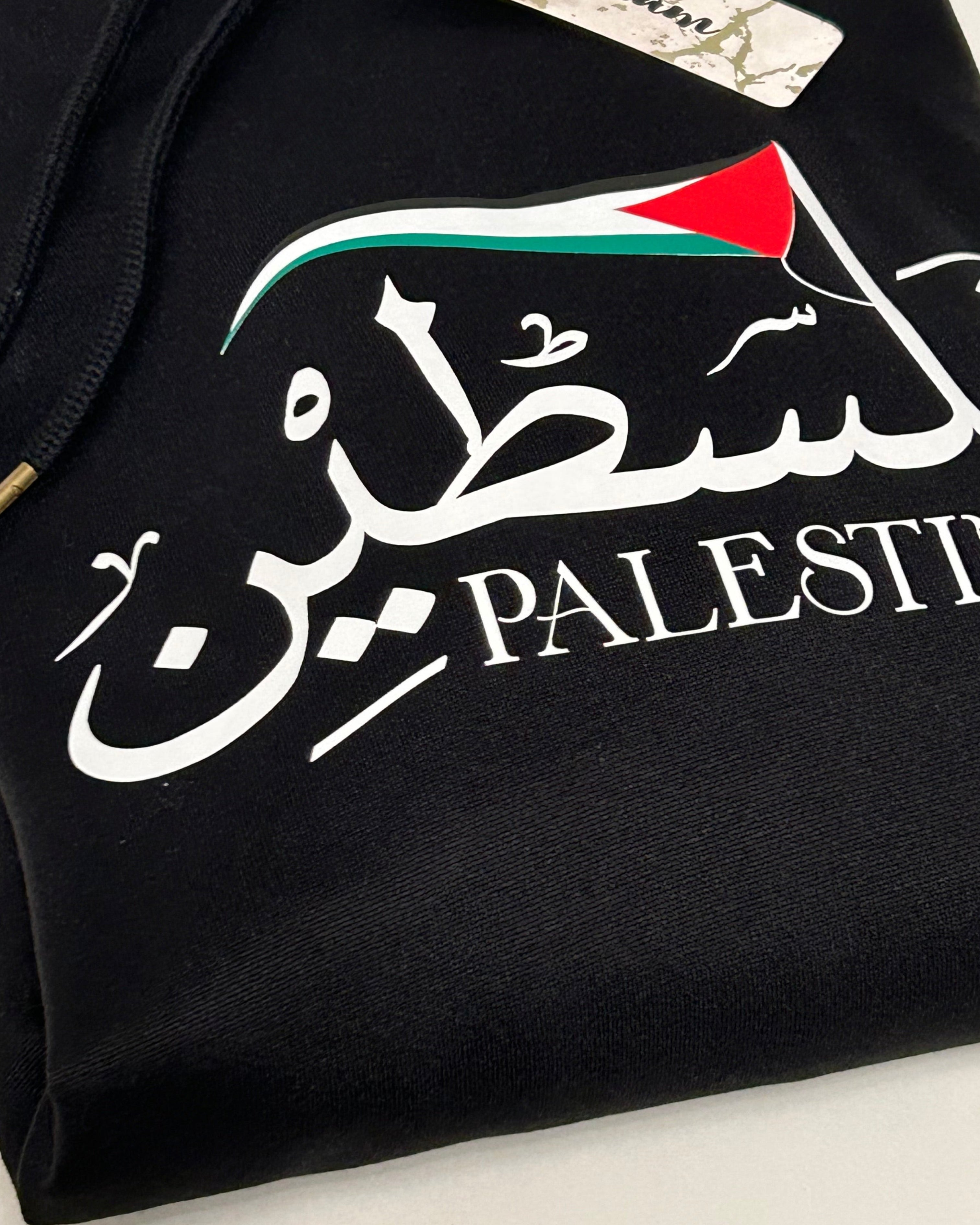 Black hoodie with Arabic text 'Palestine' and a red, black, green, and white flag on a light background