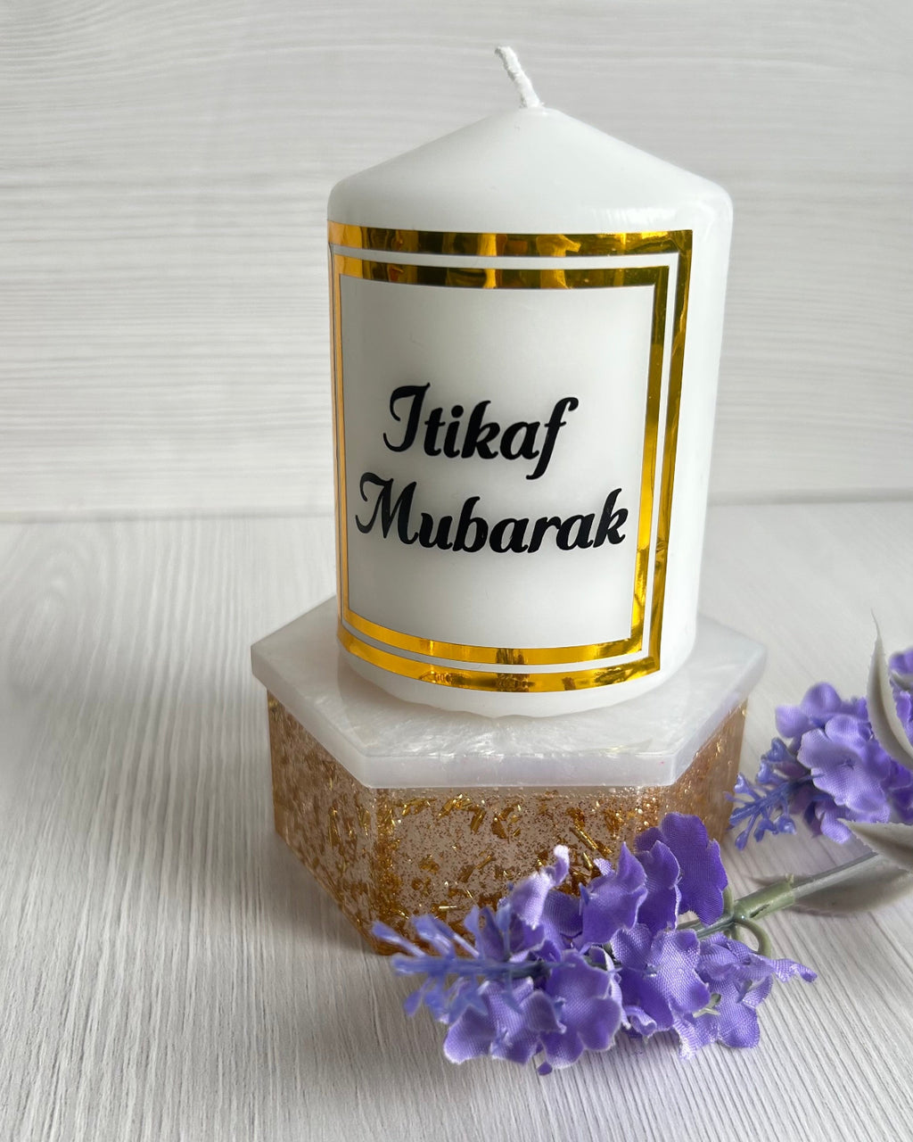 A candle personalised with Itikaf Mubarak in black text and a square gold frame around the text displayed on a gold and white display block next to purple flowers.