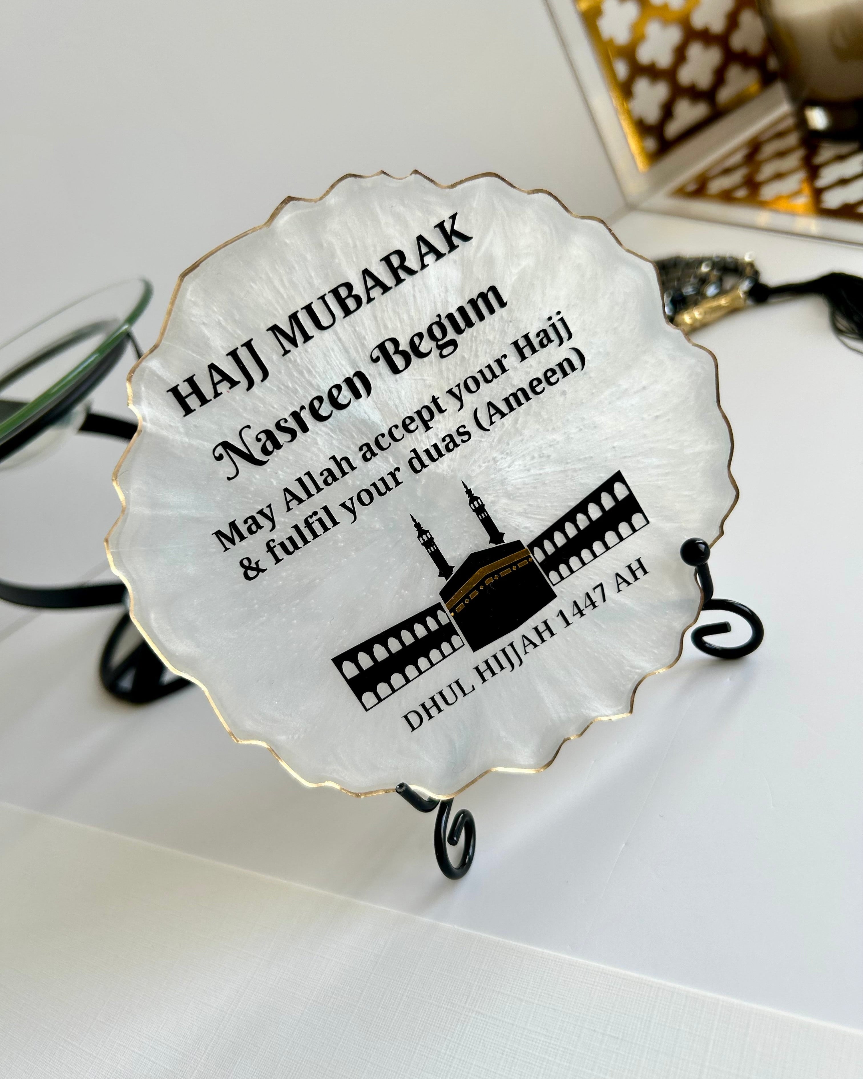 Pearlescent decorative Hajj Mubarak Plaque with text and the Kaaba and Mosque silhouette on a black display stand on a white background with decorative items