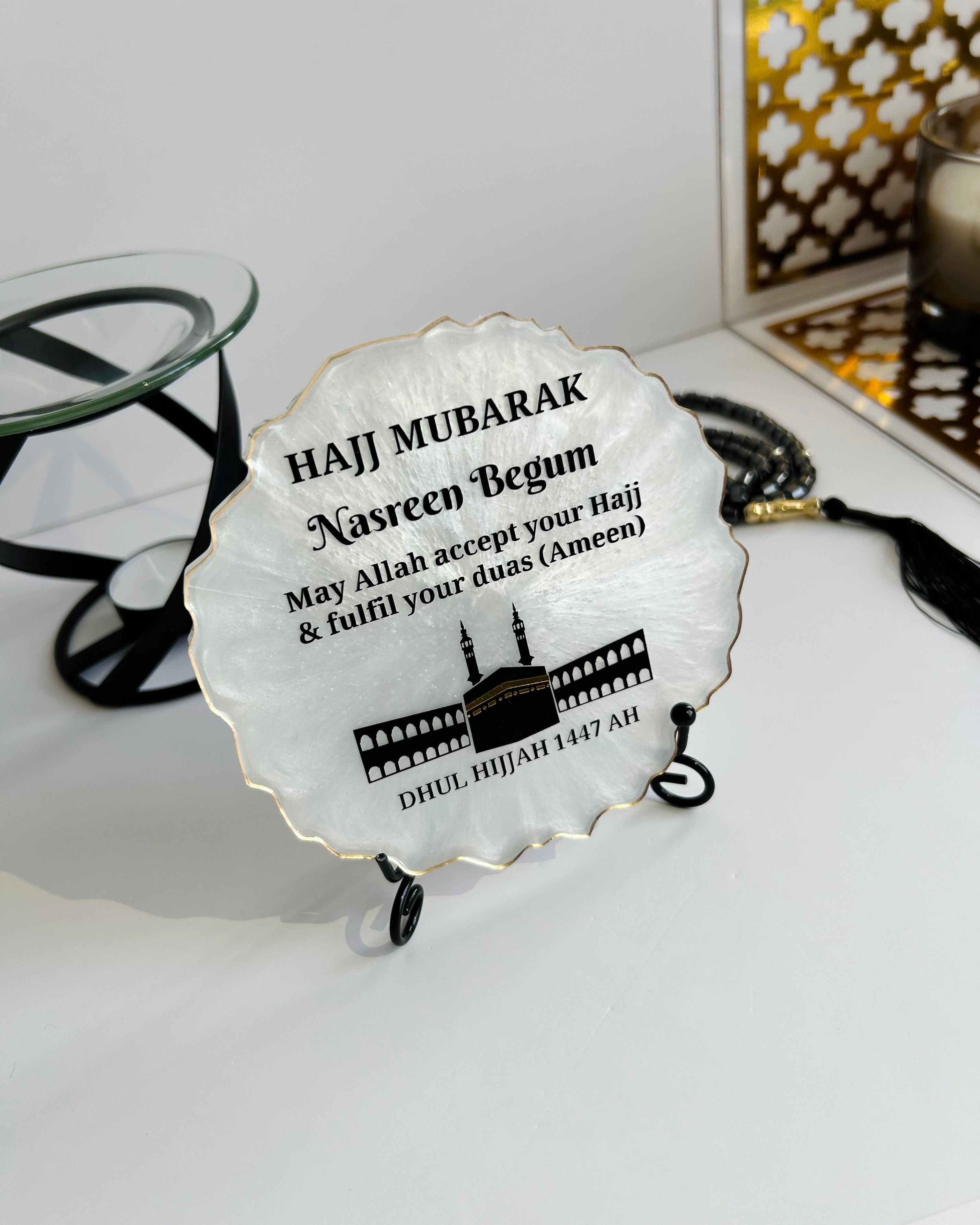 Pearlescent decorative Hajj Mubarak Plaque with text and the Kaaba and Mosque silhouette on a black display stand next to prayer beads on a white background with decorative items