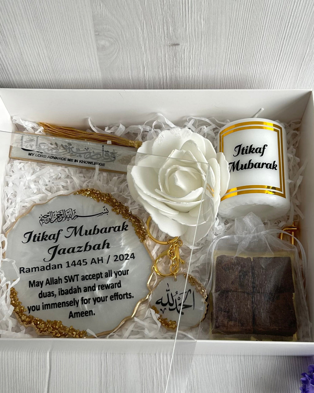A resin plaque with gold crushed glass detailing, personalized with 'Itikaf Mubarak' and some Arabic text, accompanied by a candle and a keychain with matching design, displayed alongside purple flowers.
