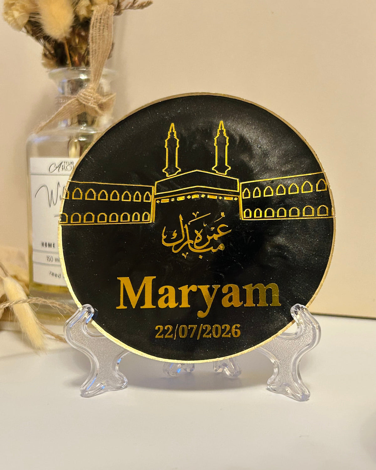 A round black plaque with gold Islamic artwork and personalised with the name 'Maryam' in gold, displayed on a clear acrylic stand on a white surface with a beige background.
