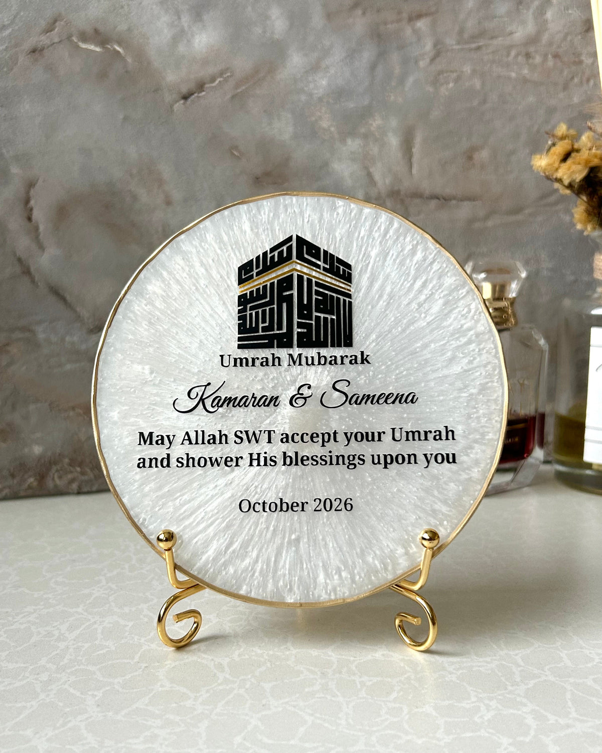 Personalised round pearlescent white Islamic plaque with gold edging displayed on a small gold display stand, featuring a black Kufic Kaaba design reading Basmala and custom text for Umrah Mubarak, on a light marble surface.