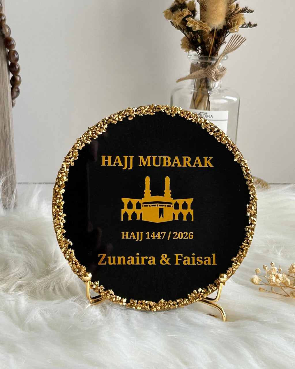 Round black Hajj Mubarak plaque with gold rim and text on a white surface with decorative elements.