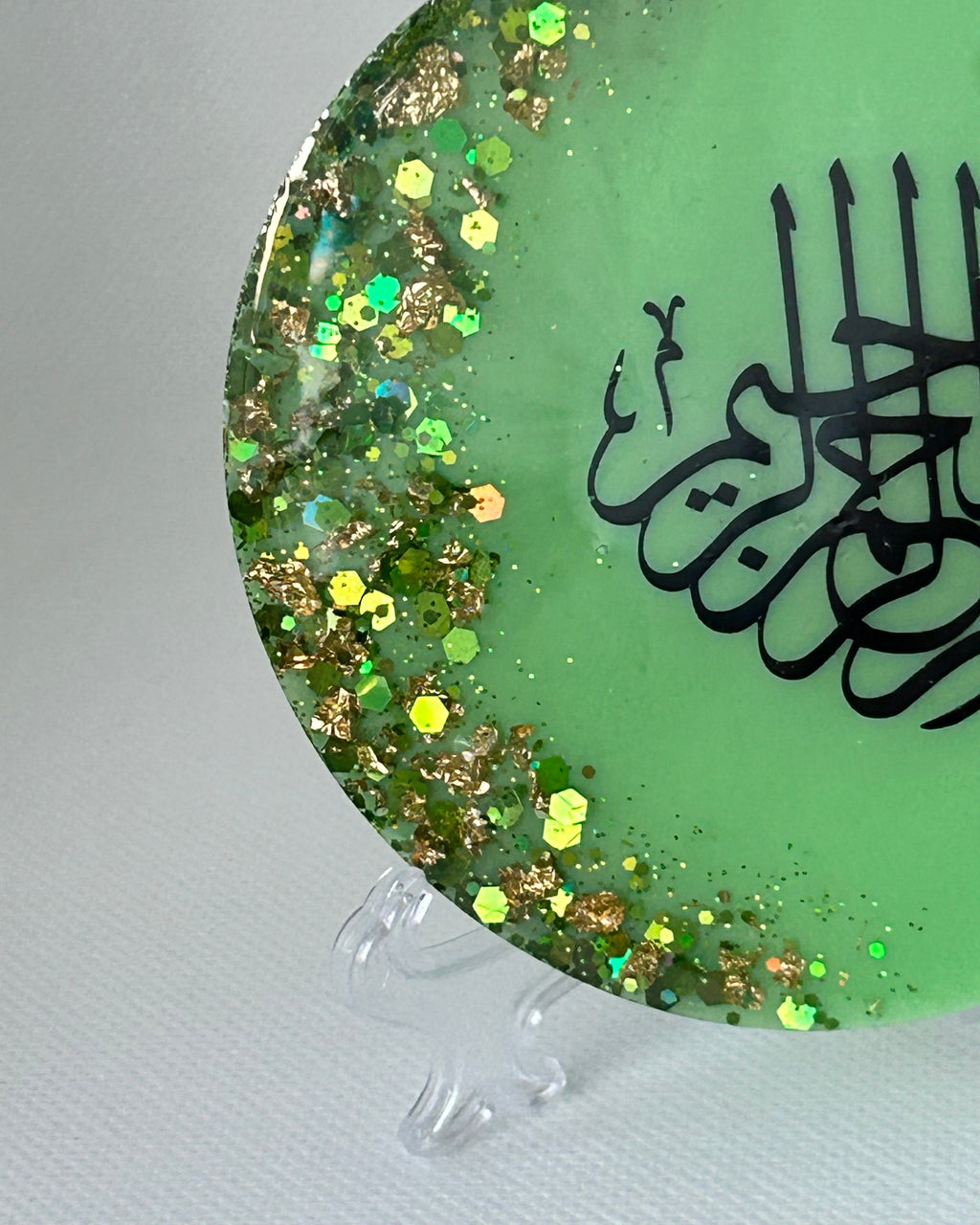Partial Green circular object with black Arabic calligraphy reading Bismillah Tasmiyah and gold shimmer glitter with gold leaf foiled placed sparsely displayed on a clear plastic display stand on a white background