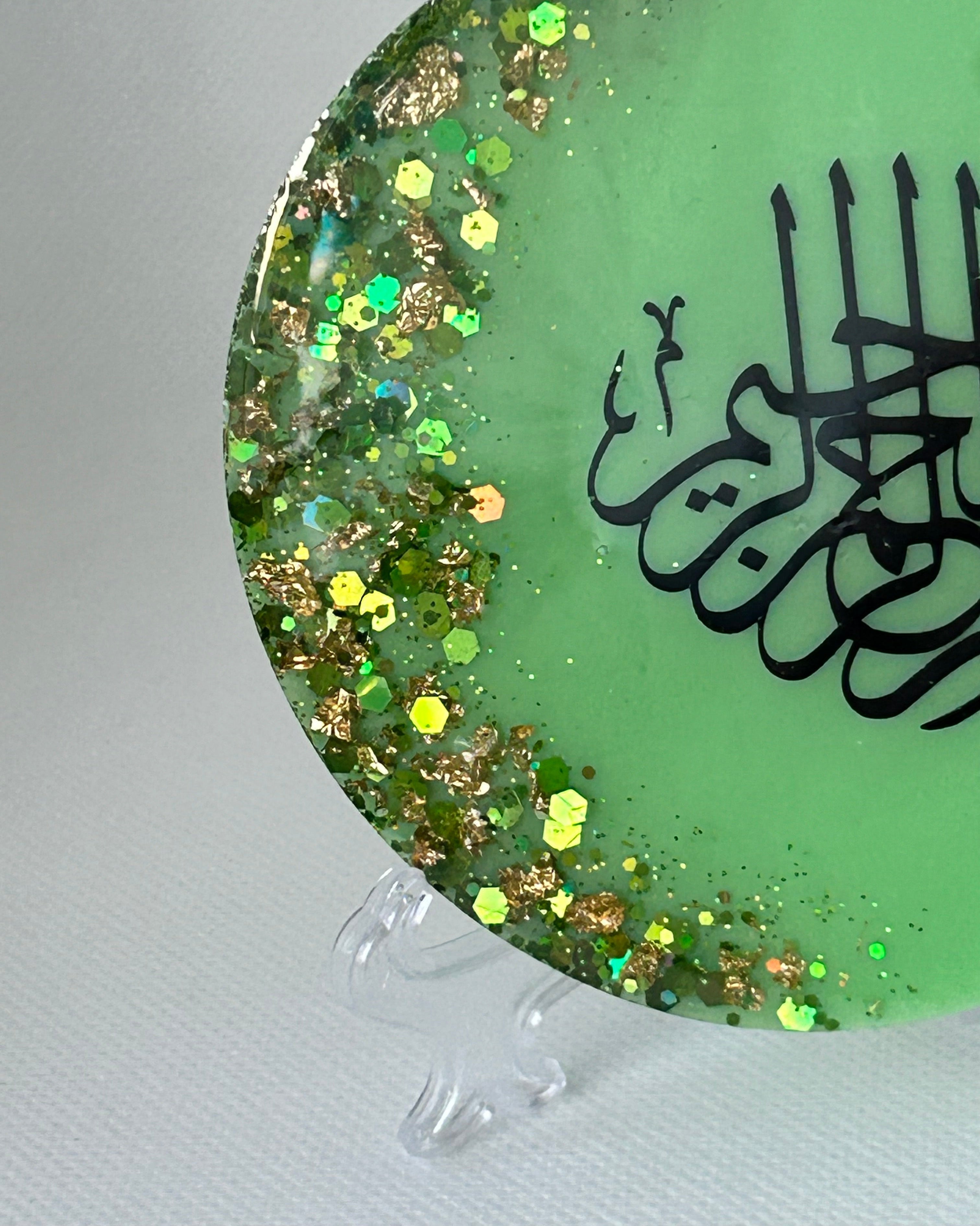 Partial Green circular object with black Arabic calligraphy reading Bismillah Tasmiyah and gold shimmer glitter with gold leaf foiled placed sparsely displayed on a clear plastic display stand on a white background