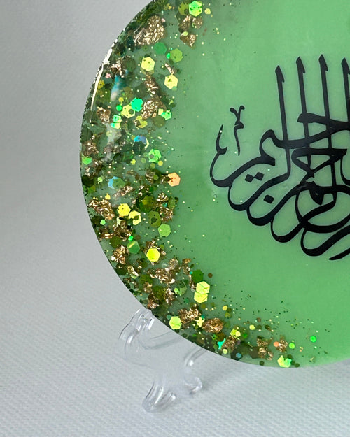Partial Green circular object with black Arabic calligraphy reading Bismillah Tasmiyah and gold shimmer glitter with gold leaf foiled placed sparsely displayed on a clear plastic display stand on a white background