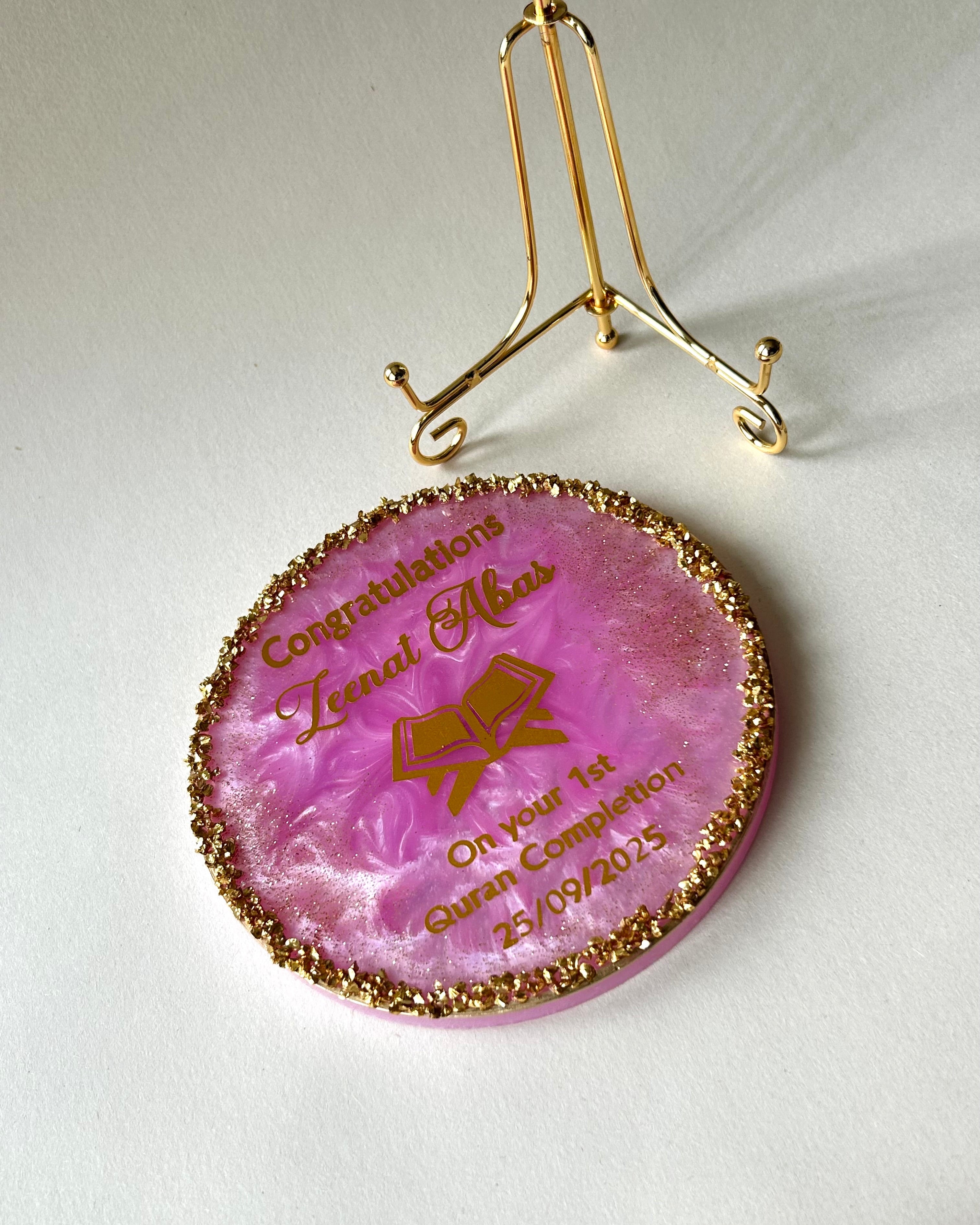 Pink decorative Quran completion plaque with gold text and design placed flat next to a gold display stand on a light textured background 