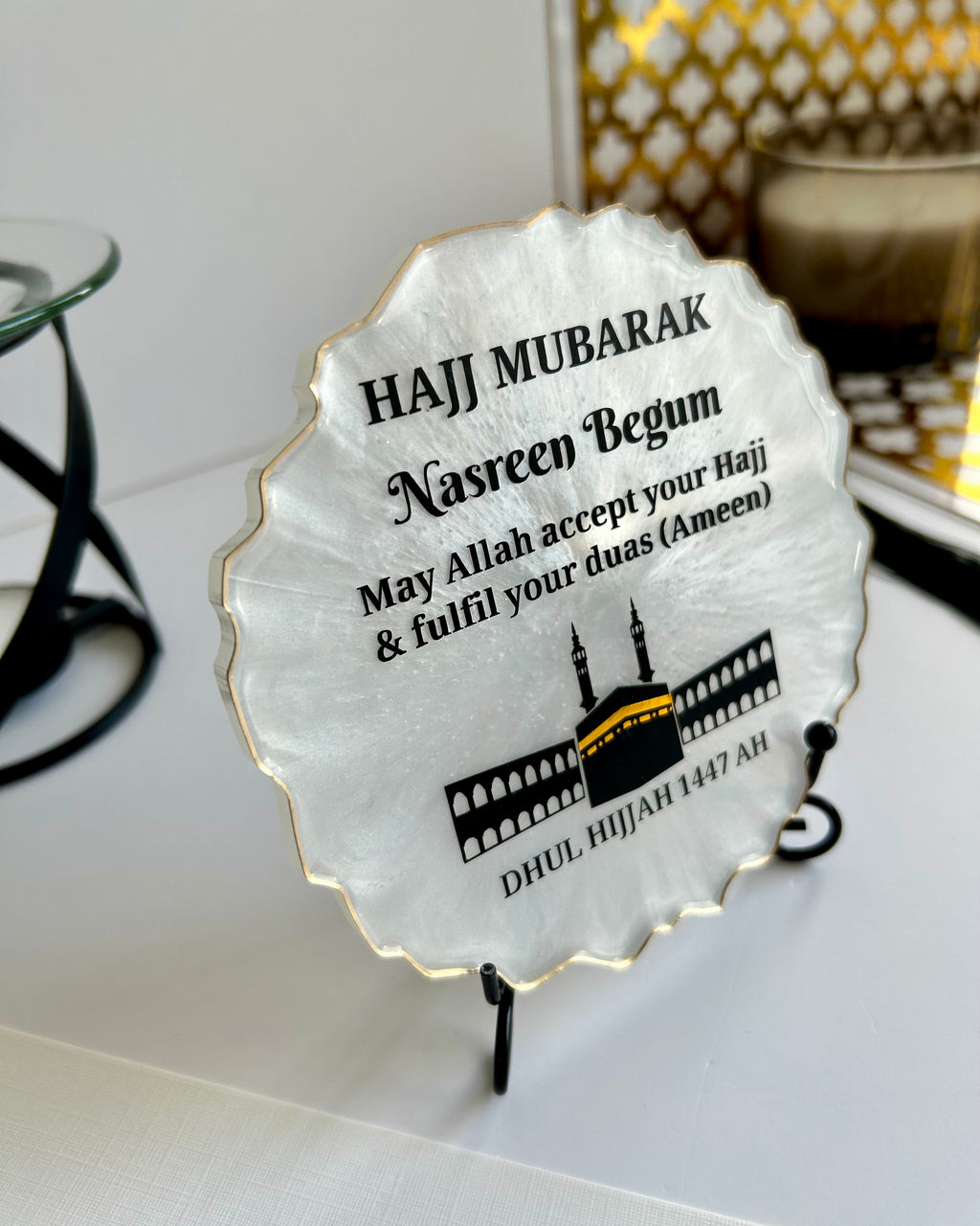 Pearlescent decorative Hajj Mubarak Plaque with text and the Kaaba and Mosque silhouette on a black display stand on a white background with decorative items