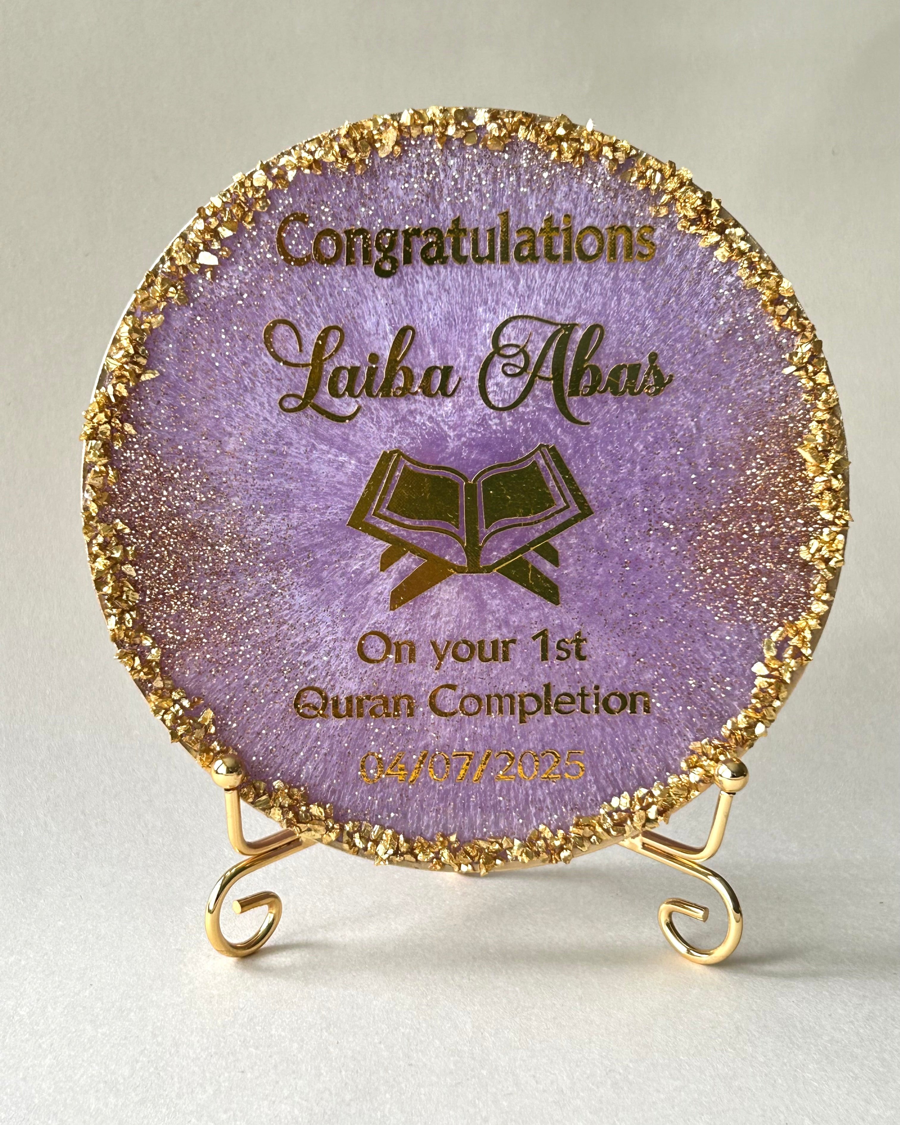Decorative Quran completion plaque with gold text and a Quran icon resting on a gold metal display stand on a white background