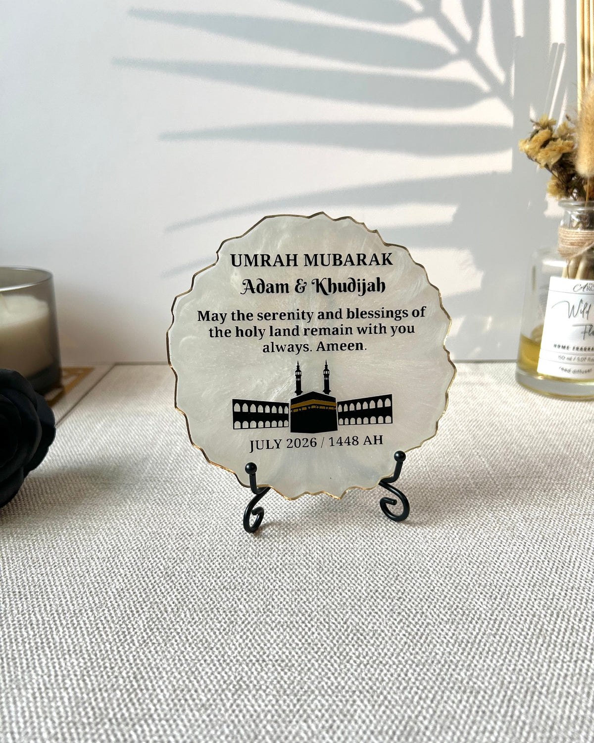 A round, scalloped-edge resin plaque with a white background and personalised in black text celebrating Umrah Mubarak, displayed on a small black stand on a textured beige fabric surface.