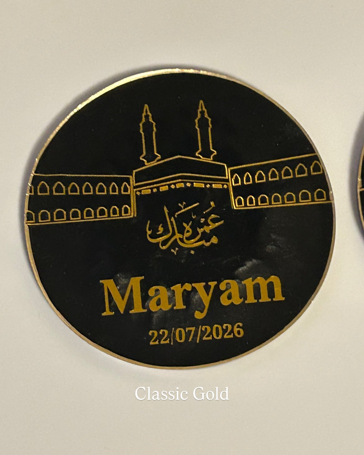 A round black and gold personalised resin Umrah Mubarak gift, featuring a gold line art illustration of the Kaaba with Arabic calligraphy and the name 'Maryam' printed in gold, along with the date '22/07/2026'.