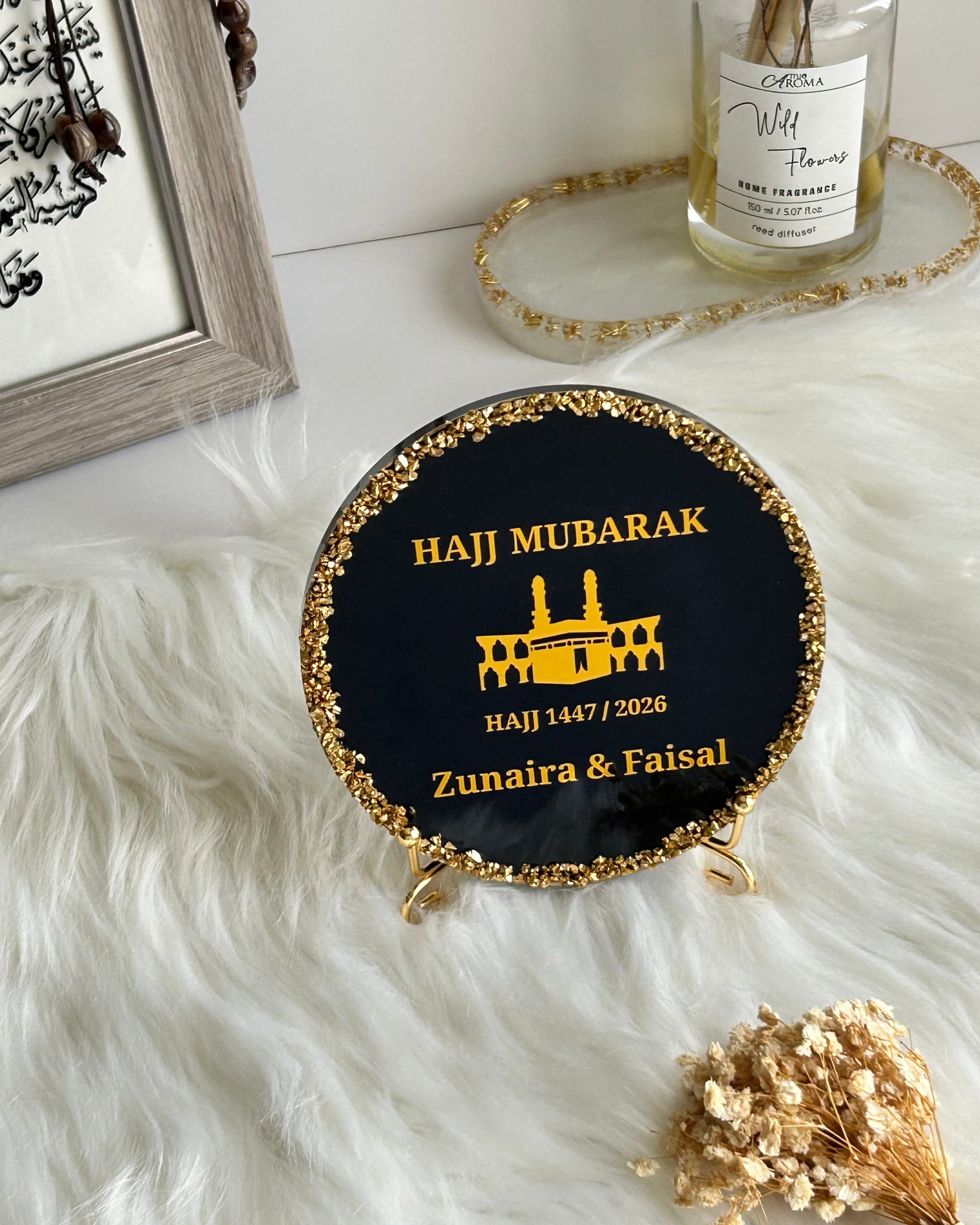 Round black decorative Hajj Mubarak plaque with gold rim and text on a white surface with decorative elements.