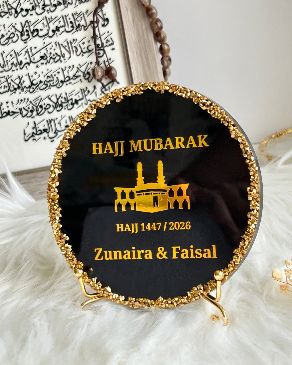 Round black Hajj Mubarak plaque with gold rim and text on a white surface with decorative elements.
