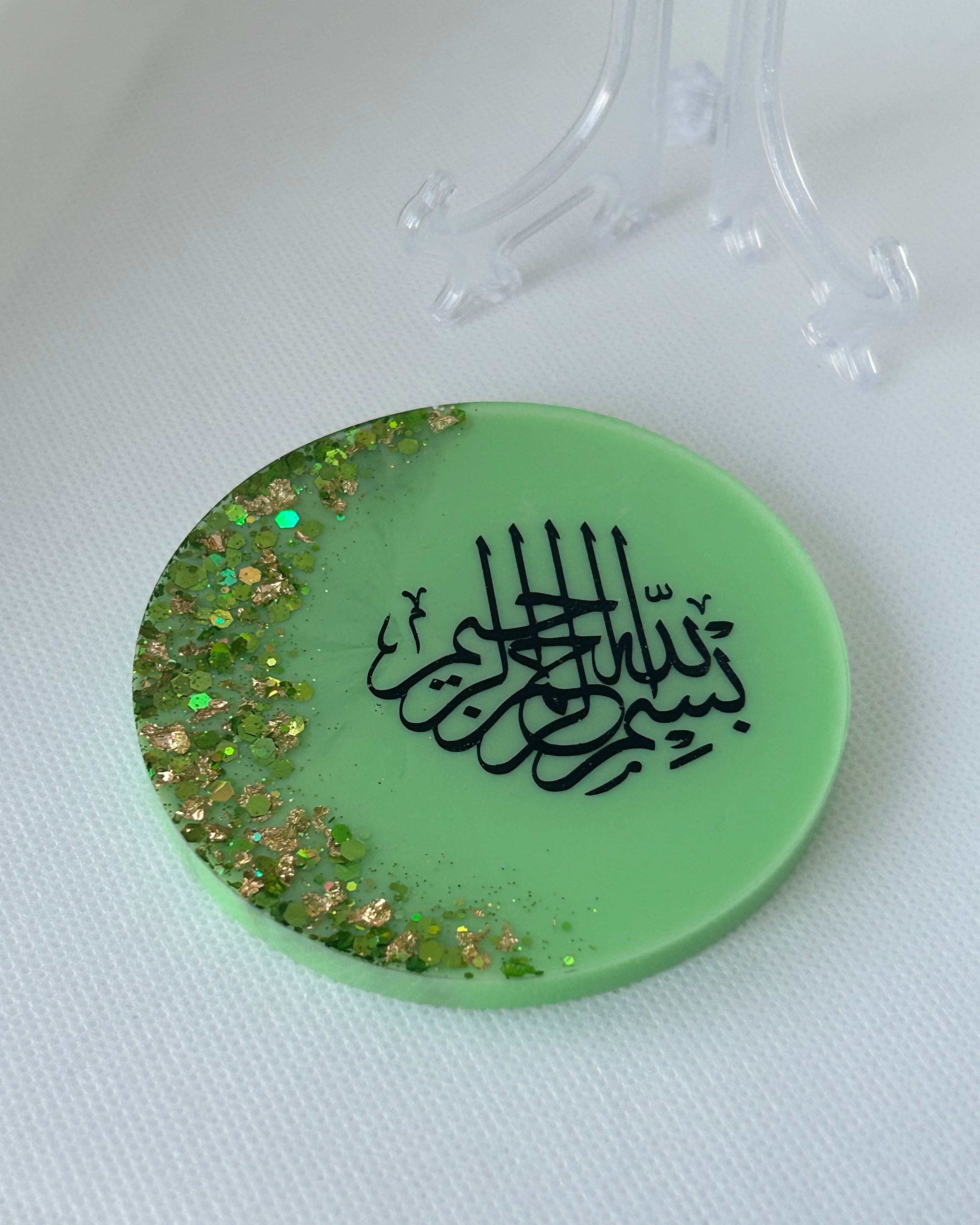 Green circular object with black Arabic calligraphy reading Bismillah Tasmiyah and gold shimmer glitter with gold leaf foiled placed sparsely displayed on a clear plastic display stand on a white background