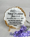 A resin plaque with gold crushed glass detailing, personalized with 'Itikaf Mubarak' and some Arabic text, accompanied by a candle and a keychain with matching design, displayed alongside purple flowers.